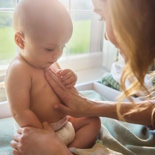 Mutter streichelt liebevoll die nackte, windeltragende Babybrust am Wickelplatz im warmen Sonnenlicht – sanfte Pflege für sensible Haut.