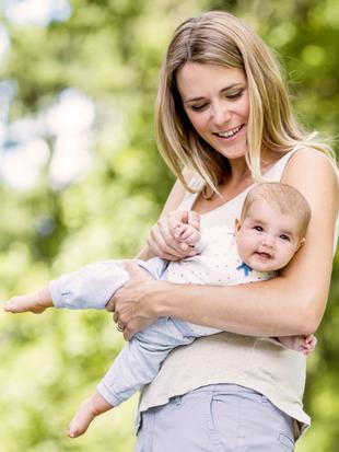 Eine Frau hält ein Baby im Freien mit Grünzeug im Hintergrund.