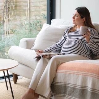 A pregnant woman relaxes on a sofa with a mug in one hand and a magazine in the other, gazing out the window. She’s dressed in a striped top and cream pants, surrounded by cozy blankets and soft natural light.