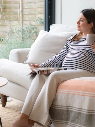 Pregnant woman relaxing on a cozy couch with a magazine and a mug, gazing out the window in a calm and peaceful setting.