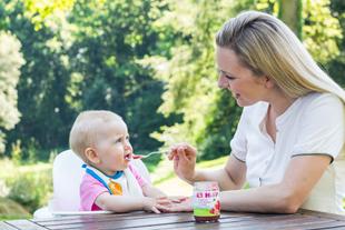 Mutter füttert ihr Baby im Hochstuhl im Garten mit einem Löffel Beikost aus einem HiPP Obstgläschen.