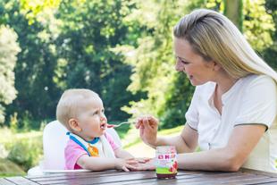 Mutter füttert ihr Baby im Hochstuhl im Garten mit einem Löffel Beikost aus einem HiPP Obstgläschen.