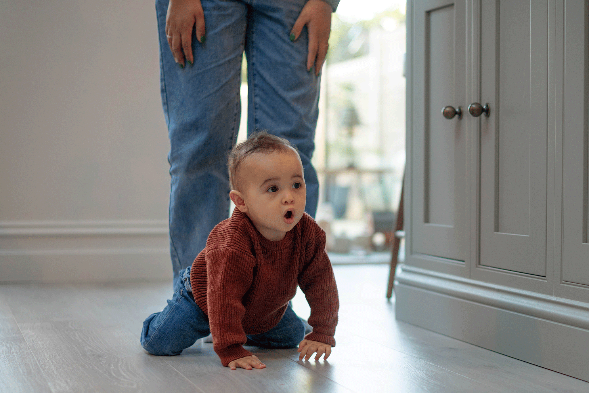 A baby wearing a rust-brown knitted jumper and jeans crawling across a light wooden floor with an adult standing closely behind.