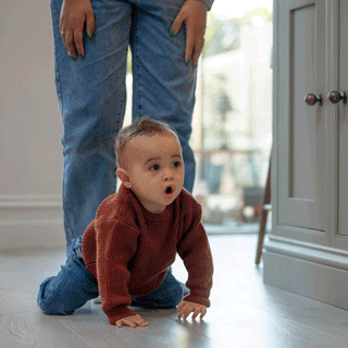 A baby wearing a rust-brown knitted jumper and jeans crawling across a light wooden floor with an adult standing closely behind.
