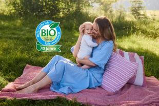 Lachende Mutter hält ihr Baby im Arm hoch und schaut es liebevoll an, in heller, natürlicher Umgebung.