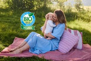 Lachende Mutter hält ihr Baby im Arm hoch und schaut es liebevoll an, in heller, natürlicher Umgebung.