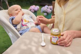 Baby sitzt in Hochstuhl und wird von Person mit hellem Hemd gefüttert, auf Tisch steht Glas mit Babybrei und Löffel