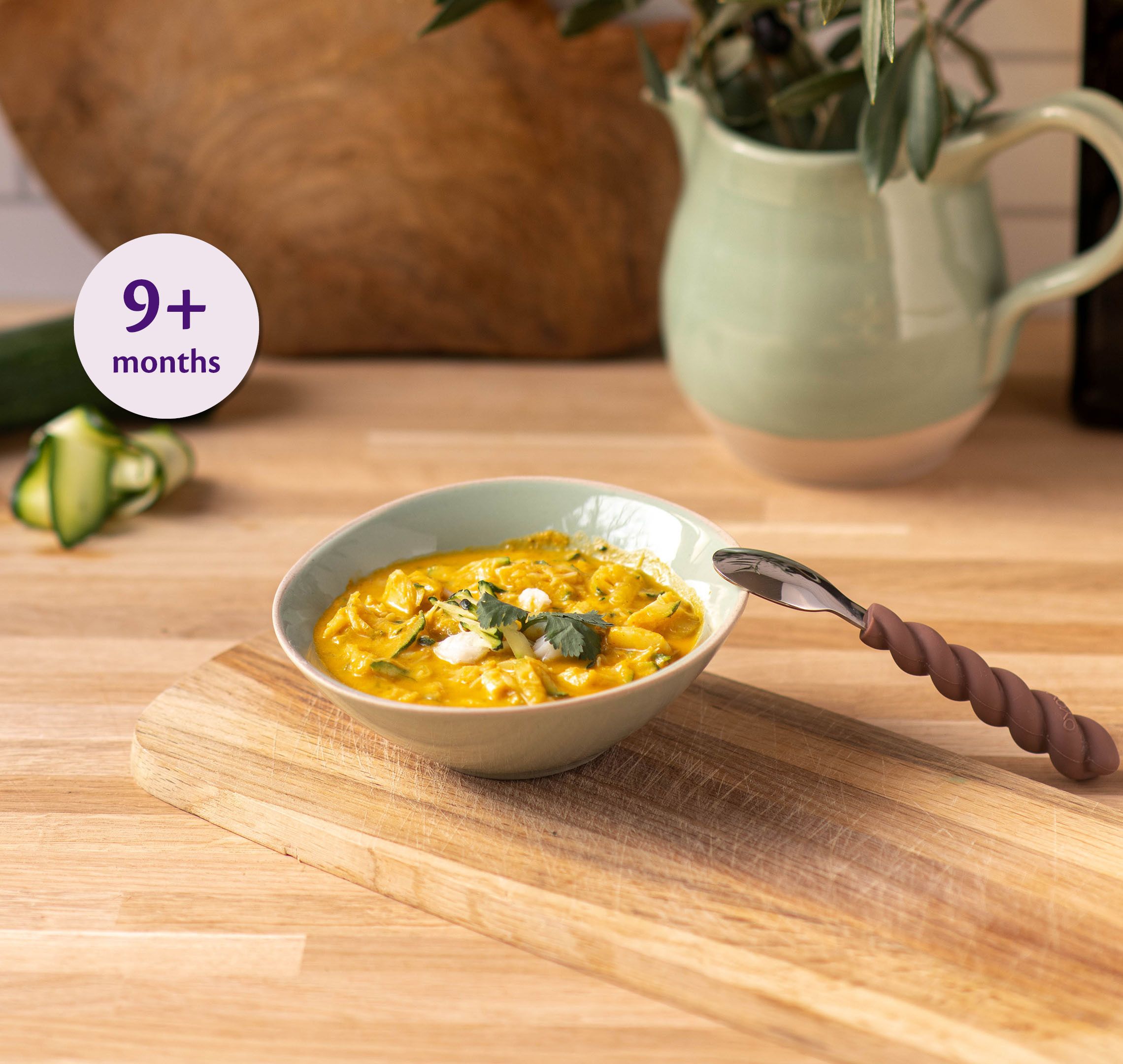 Bowl of mild fish and courgette curry for babies 9+ months on a wooden board with a baby spoon beside it
