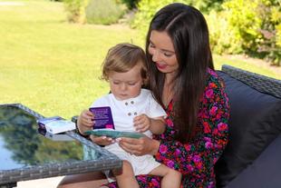 Mum and toddler sitting in a sunny garden, reading a play card together at an outdoor table