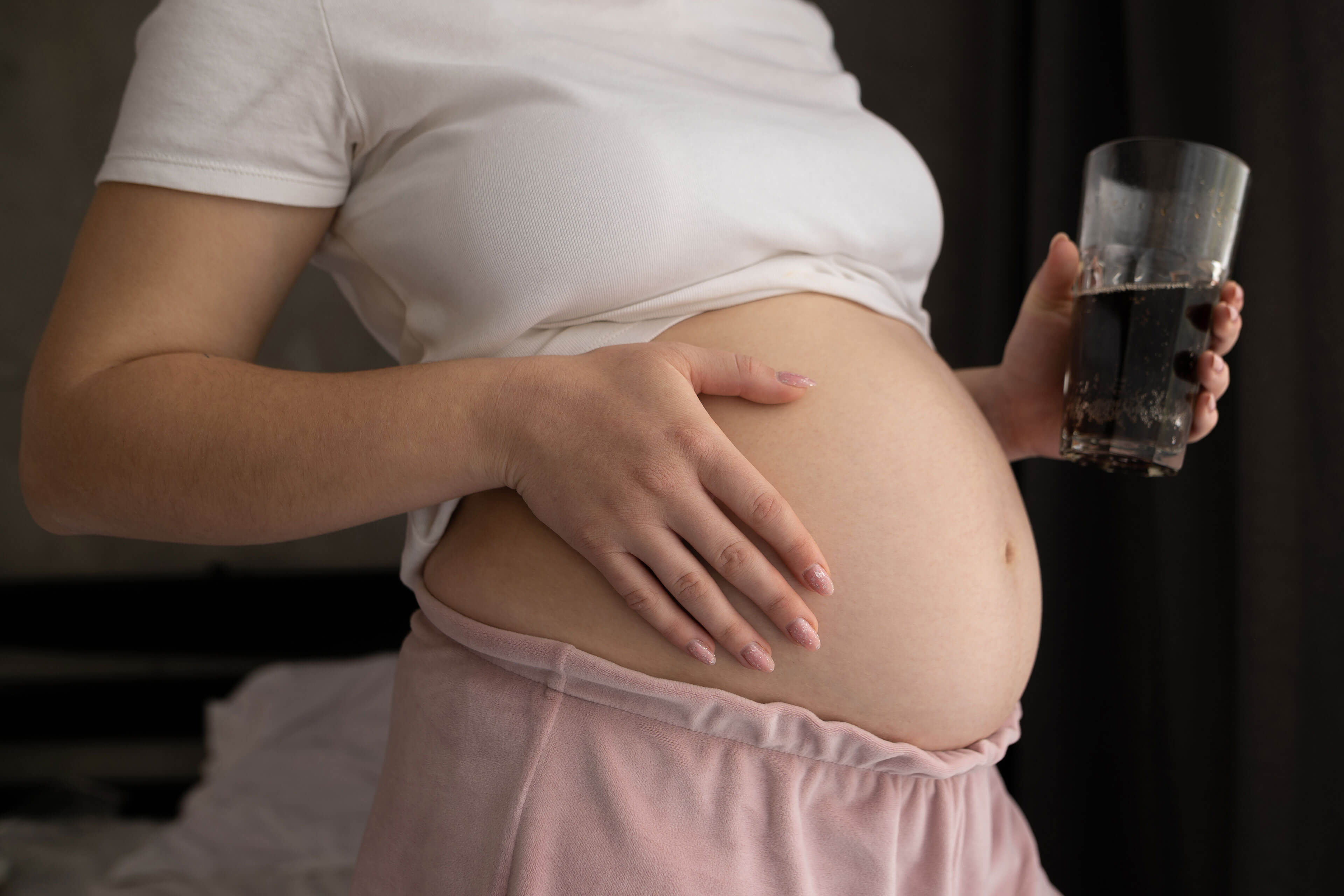 Eine Person, die ein Glas mit einer dunklen Flüssigkeit in der Hand hält, die Hand auf den Bauch legt und ein weißes Hemd trägt.