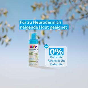 Flasche HiPP Schaumdusche mit Text 'Für zu Neurodermitis neigende Haut geeignet' und Hinweis '0% Duftstoffe Ätherische Öle Farbstoffe' vor unscharfem Hintergrund mit Pflanzenzweigen
