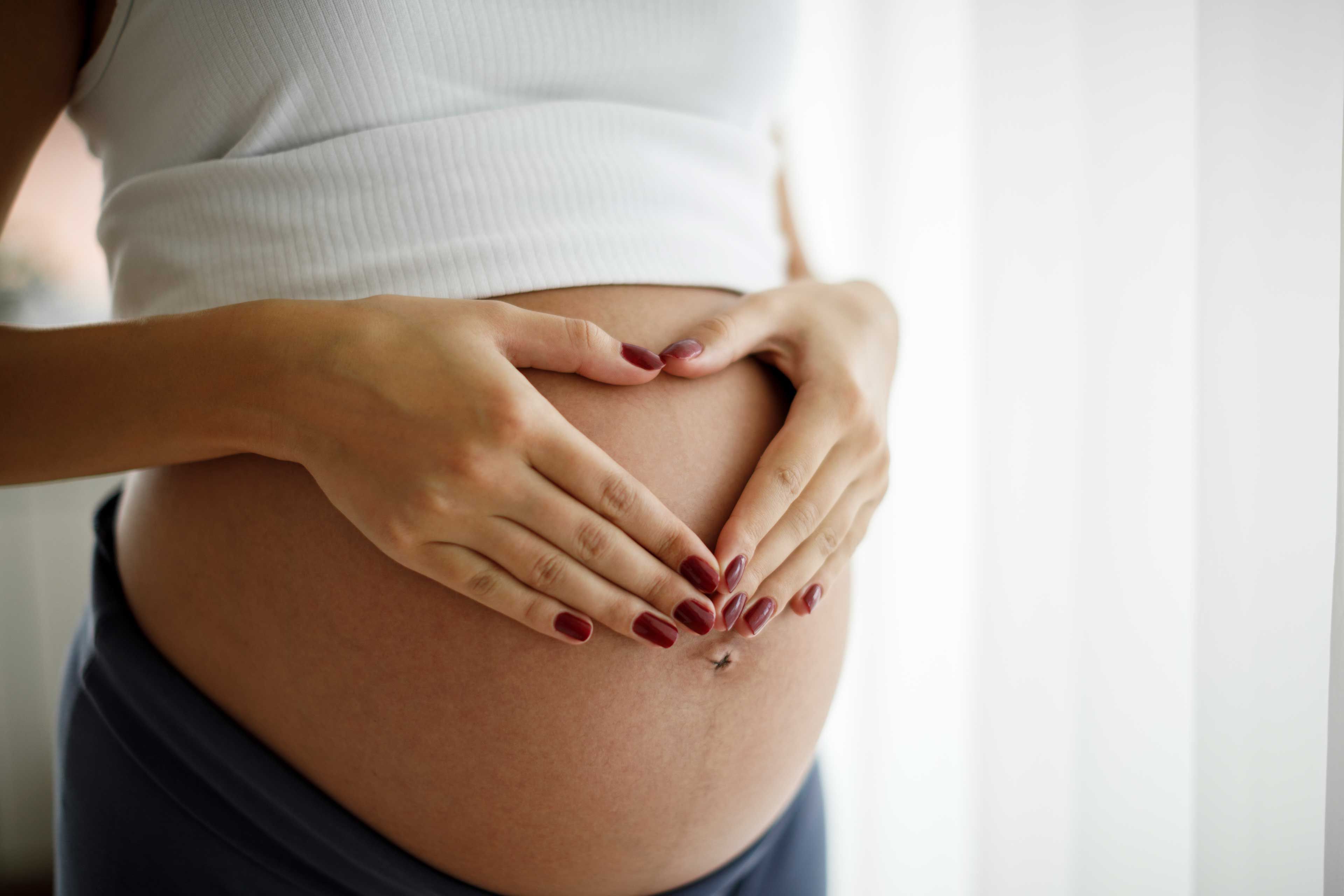 Die Hände einer Person bilden eine Herzform über dem schwangeren Bauch.