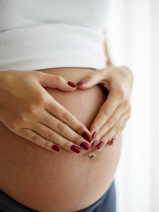 Die Hände einer Person bilden eine Herzform über dem schwangeren Bauch.