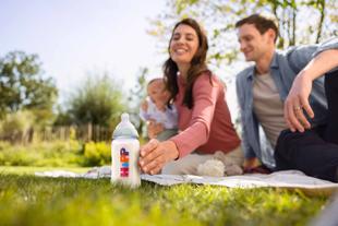 Familie mit Baby sitzt auf einer Decke im Gras, im Vordergrund steht eine Babyflasche