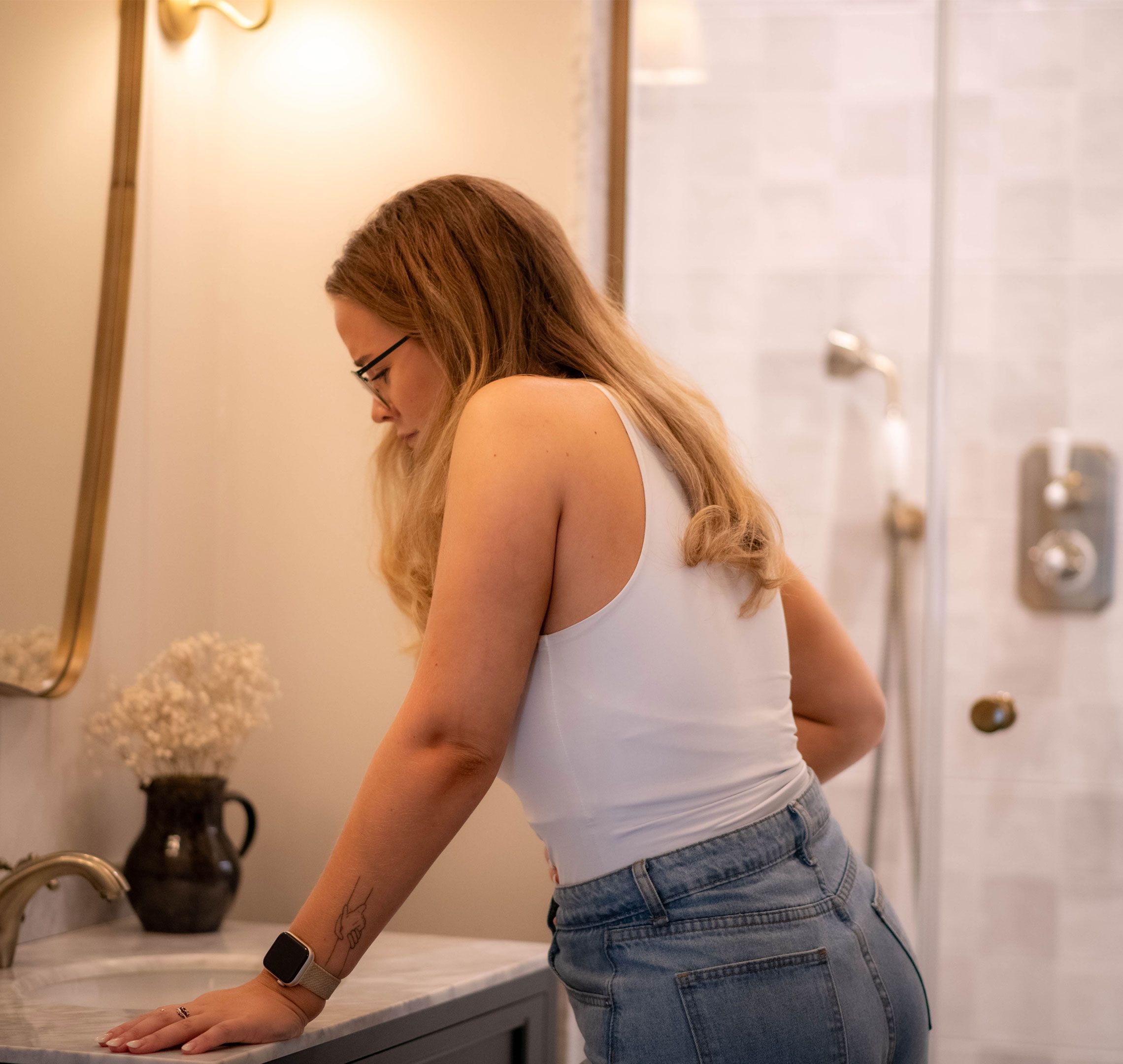 Woman in a bathroom leaning on the sink, looking down thoughtfully, possibly noticing early pregnancy spotting
