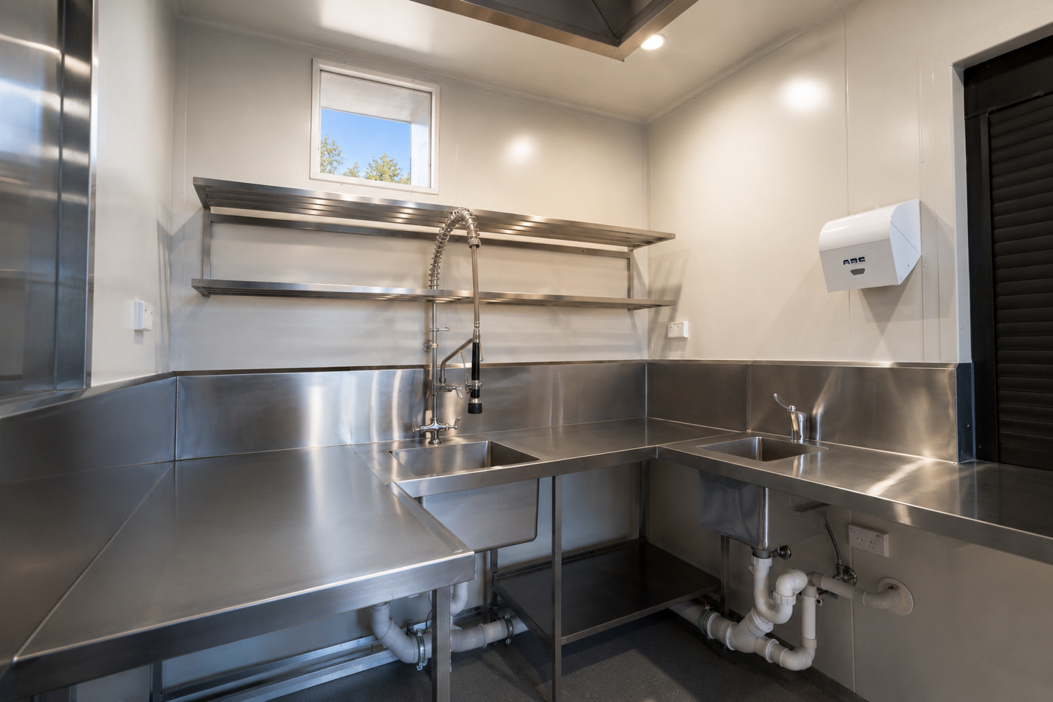 Interior of a commercial shipping container kitchen featuring stainless steel benches, double sinks, wall shelving, and a commercial pre-rinse spray unit.