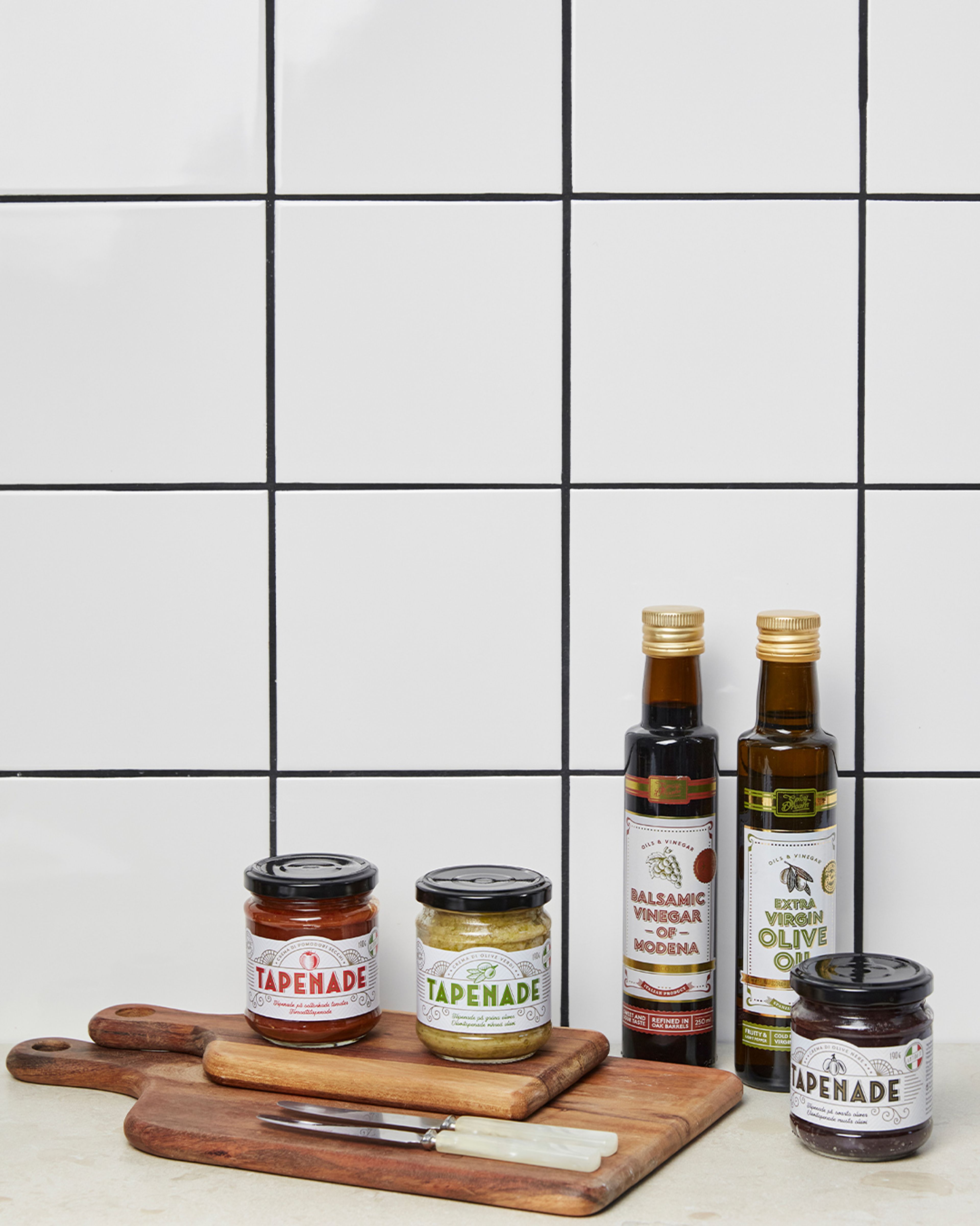Tre burkar tapenade i olika smaker tillsammans med en flaska balsamvinäger och en flaska extra jungfruolivolja, alla placerade på träskärbrädor mot en vit kakelvägg. Två små knivar med ljusa handtag ligger framför