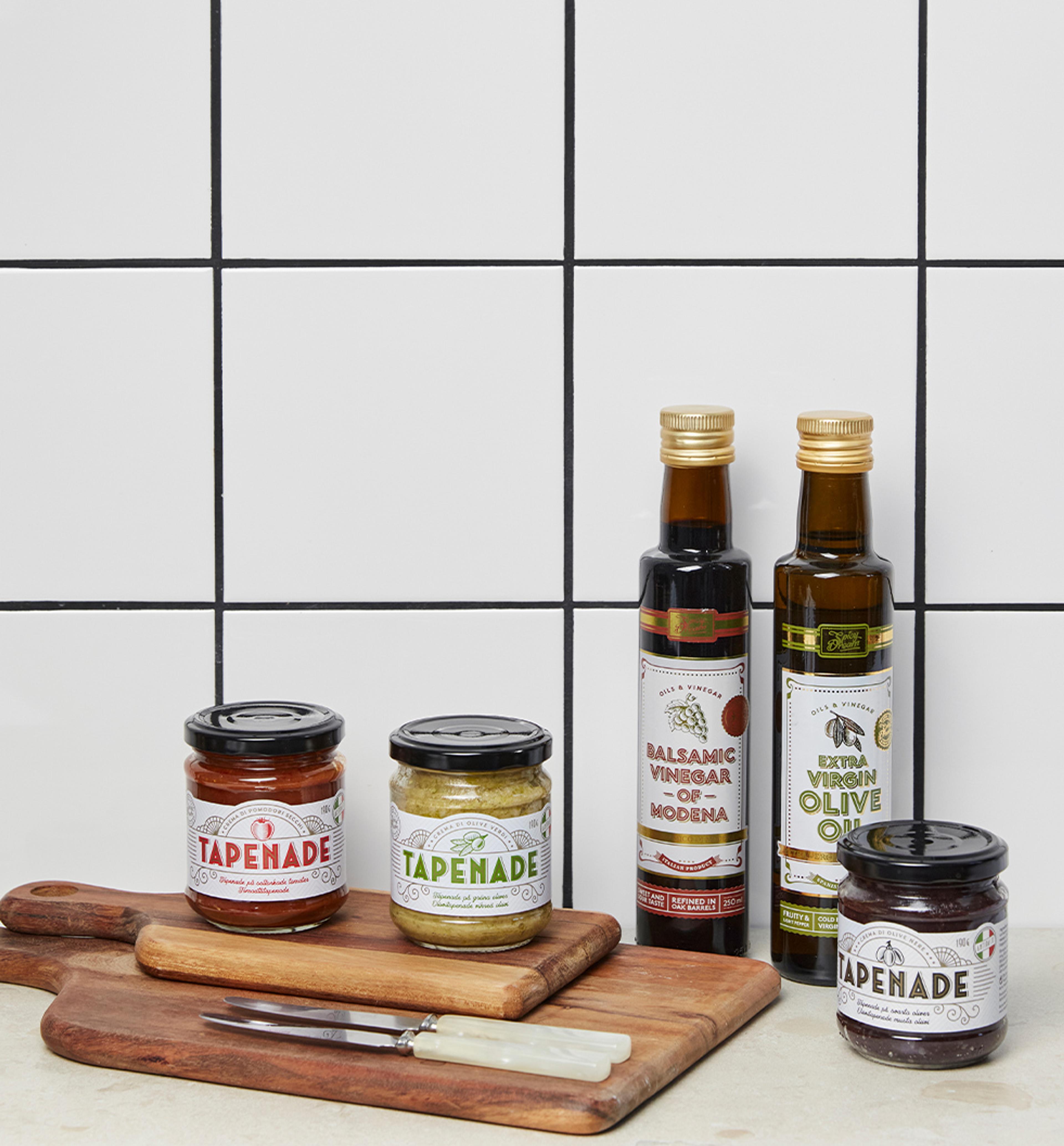 Tre burkar tapenade i olika smaker tillsammans med en flaska balsamvinäger och en flaska extra jungfruolivolja, alla placerade på träskärbrädor mot en vit kakelvägg. Två små knivar med ljusa handtag ligger framför