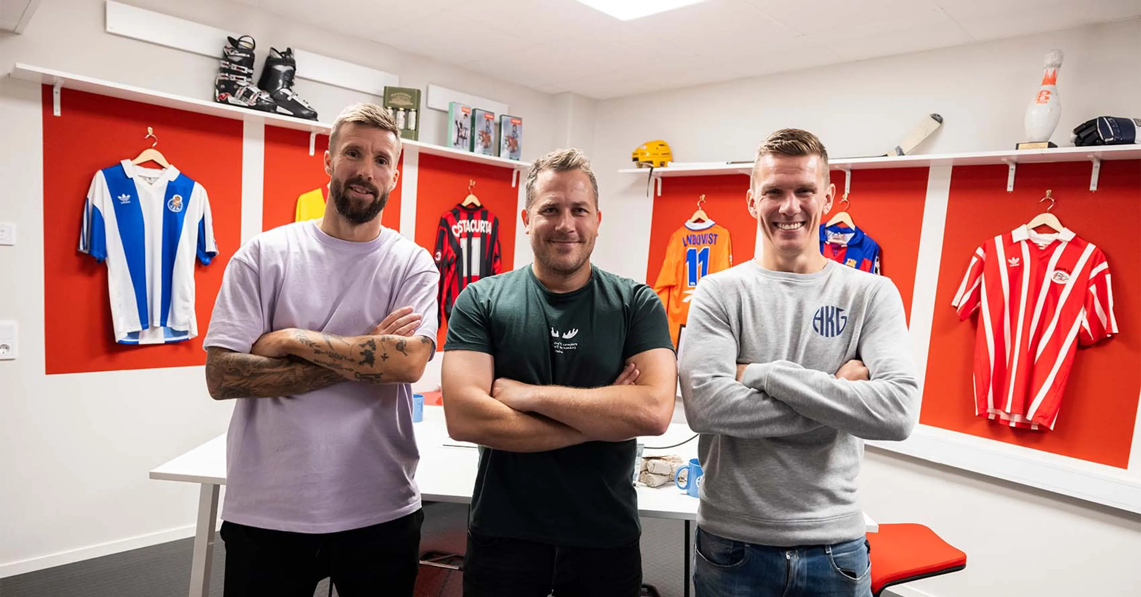 Tre män med armarna i kors står framför en vit arbetsbord i ett rum med röda väggpartier där fotbollströjor hänger uppsatta.