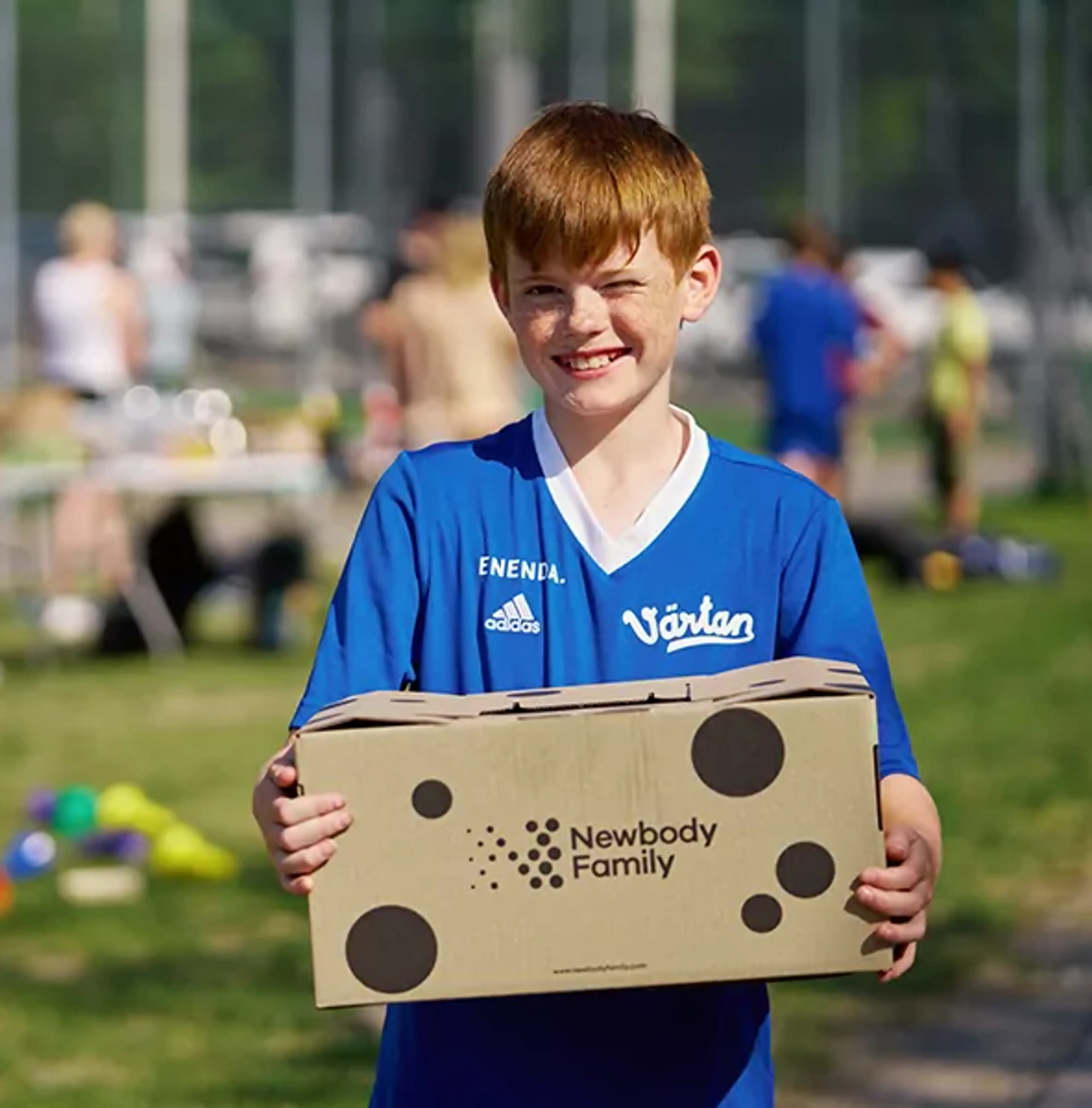 En pojke i blå fotbollströja håller en kartong med Newbody Family-logotypen på en grön gräsmatta. I bakgrunden syns flera barn och ett bord med olika saker.