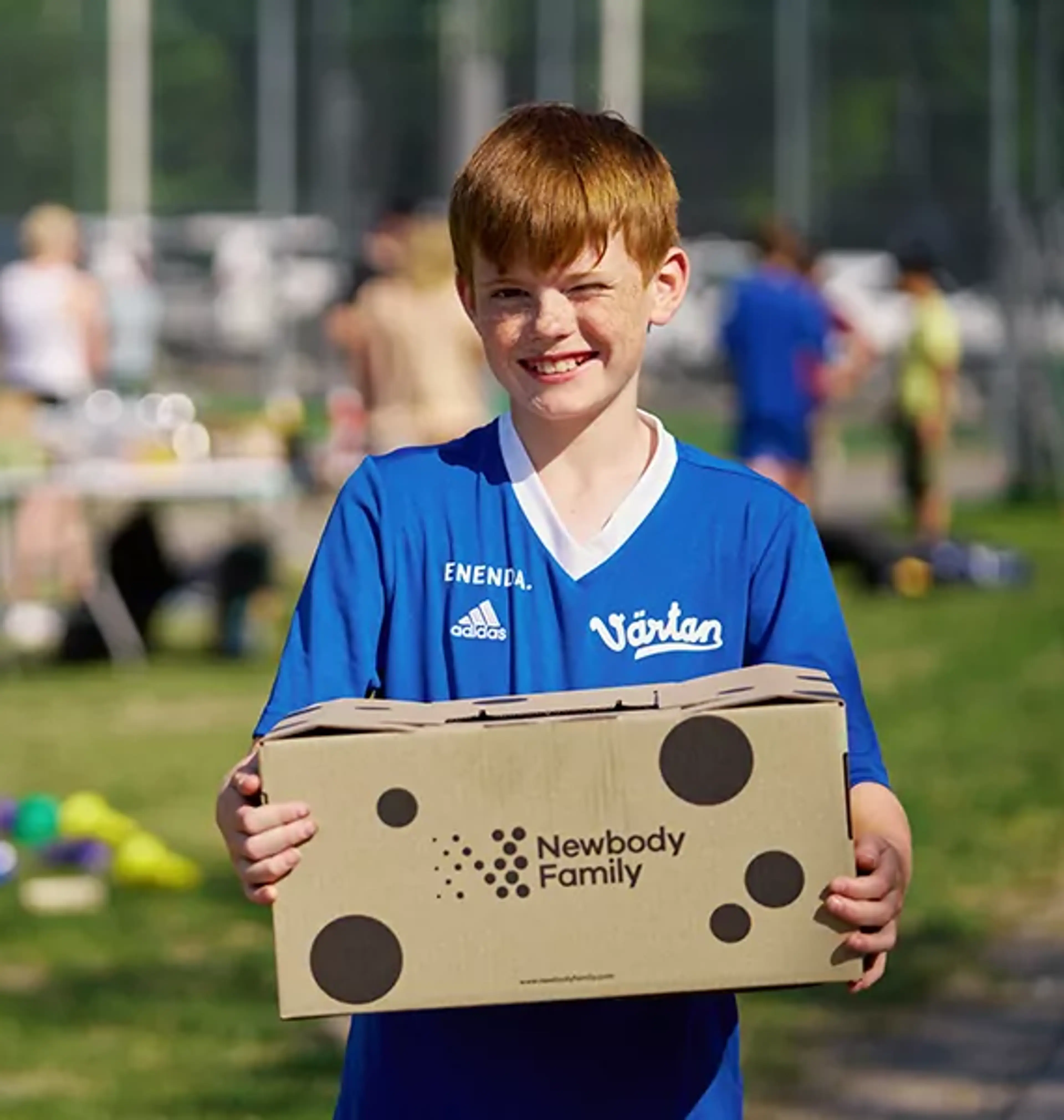 En pojke i blå fotbollströja håller en kartong med Newbody Family-logotypen på en grön gräsmatta. I bakgrunden syns flera barn och ett bord med olika saker.