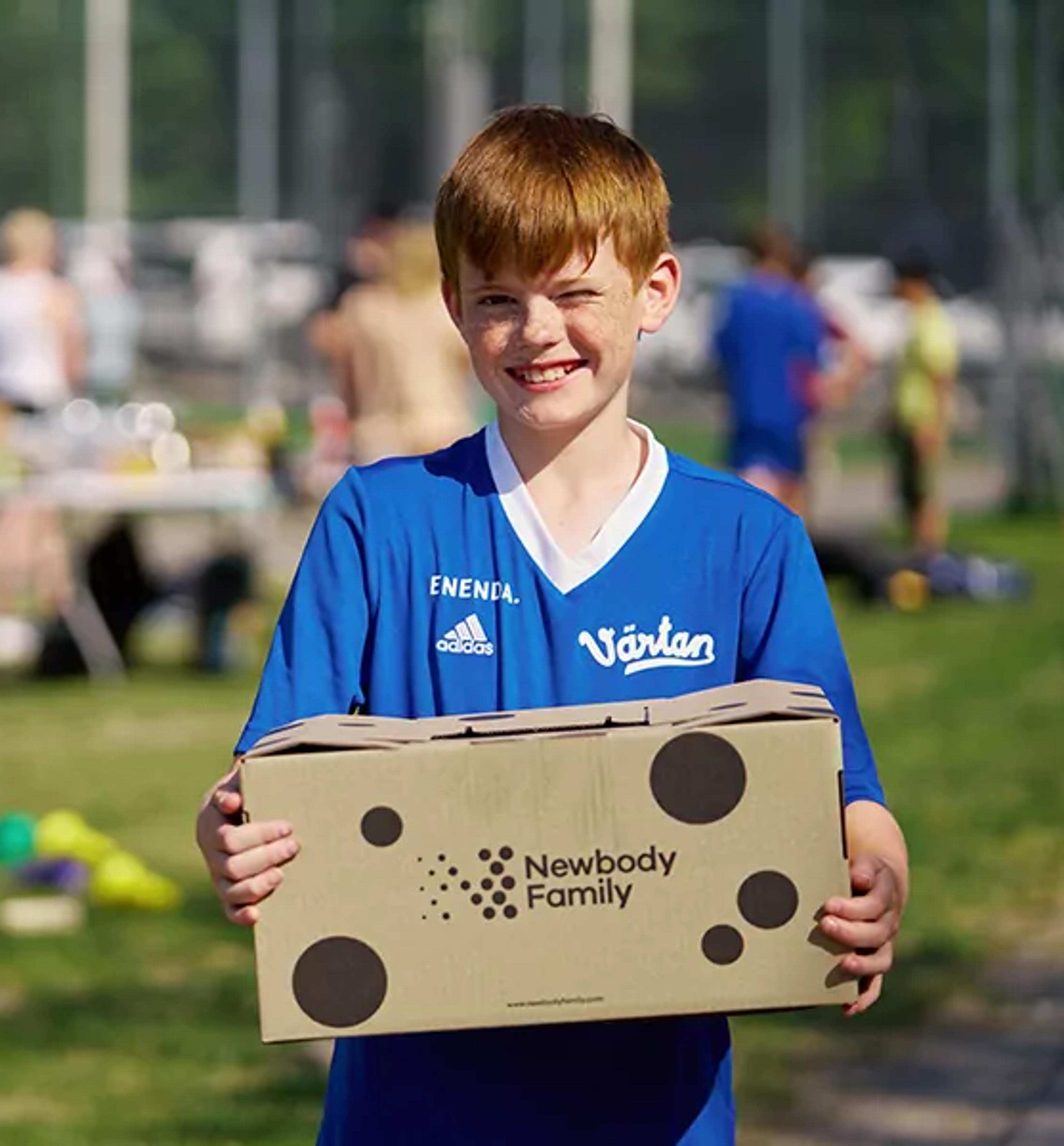 En pojke i blå fotbollströja håller en kartong med Newbody Family-logotypen på en grön gräsmatta. I bakgrunden syns flera barn och ett bord med olika saker.