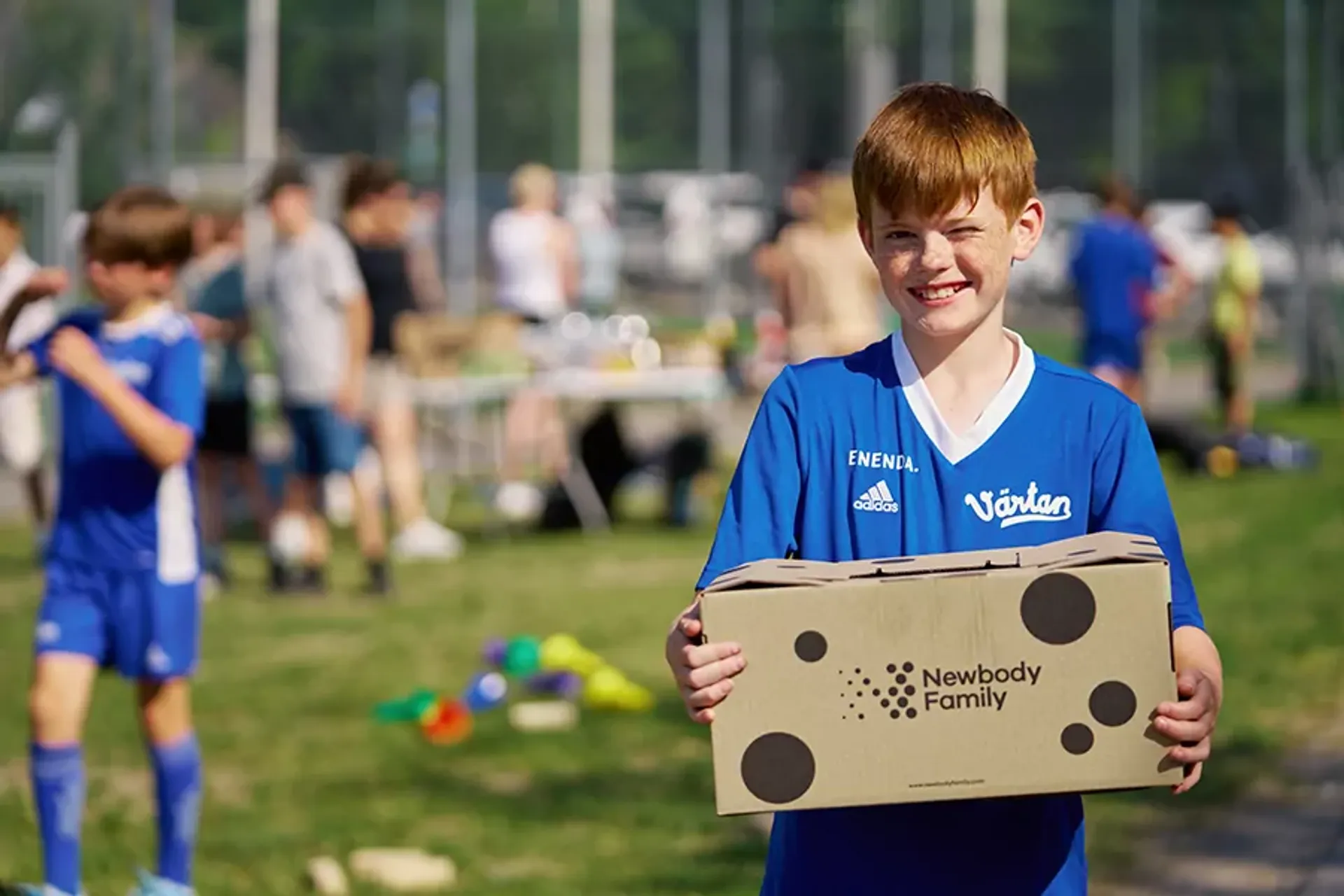 En pojke i blå fotbollströja håller en kartong med Newbody Family-logotypen på en grön gräsmatta. I bakgrunden syns flera barn och ett bord med olika saker.