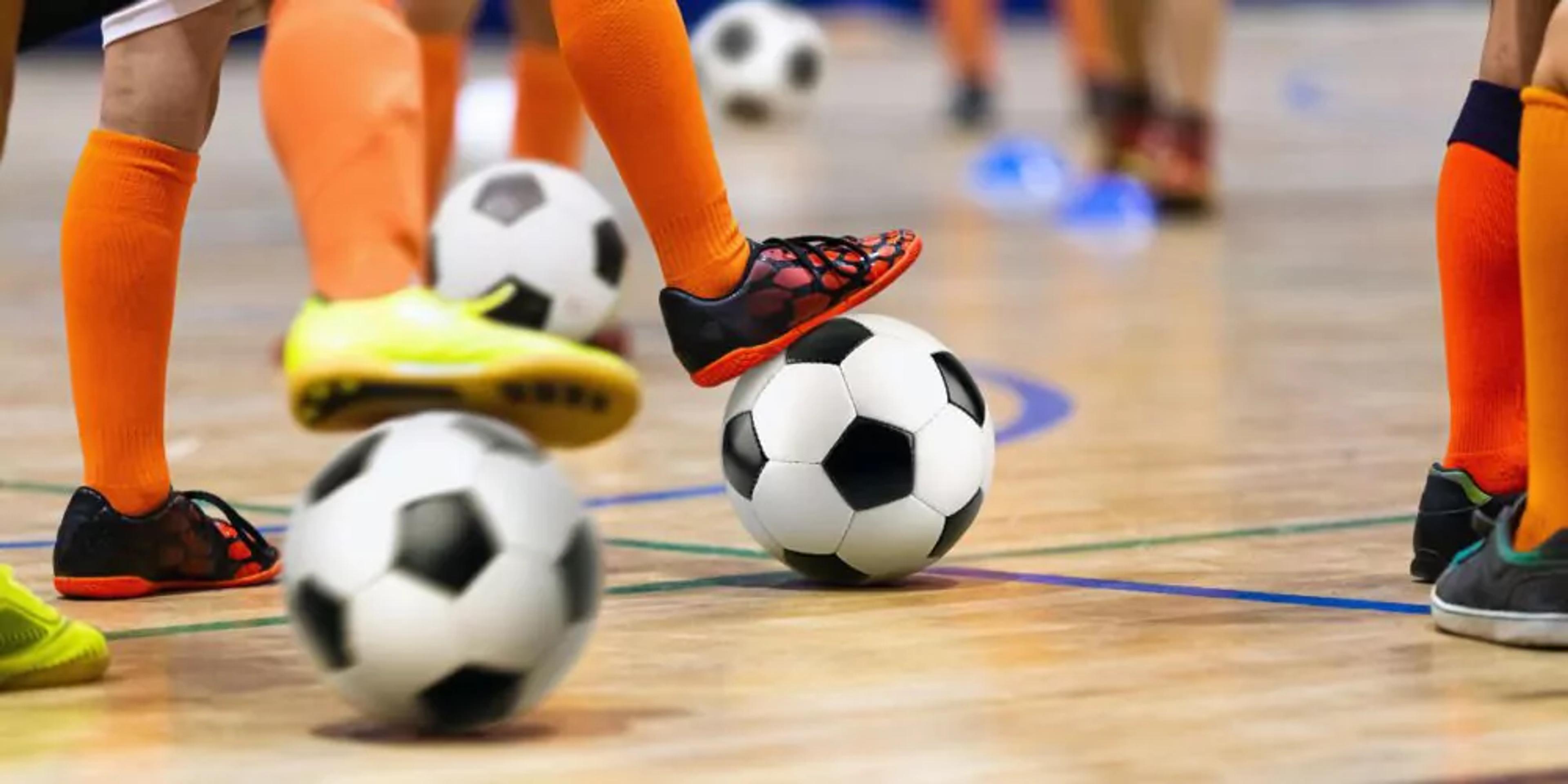 Flera barn med orange strumpor tränar futsal inomhus, där en fot är placerad på en svartvit boll. Golvet är av trä och fokuset ligger på deras fötter och bollar.