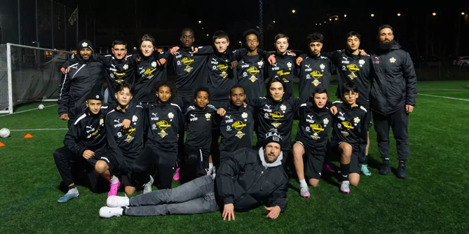 Ungdomar i matchställ står och sitter på en fotbollsplan på kvällen, två ledare är med i gruppen. Gruppen poserar samlat med armarna om varandra.