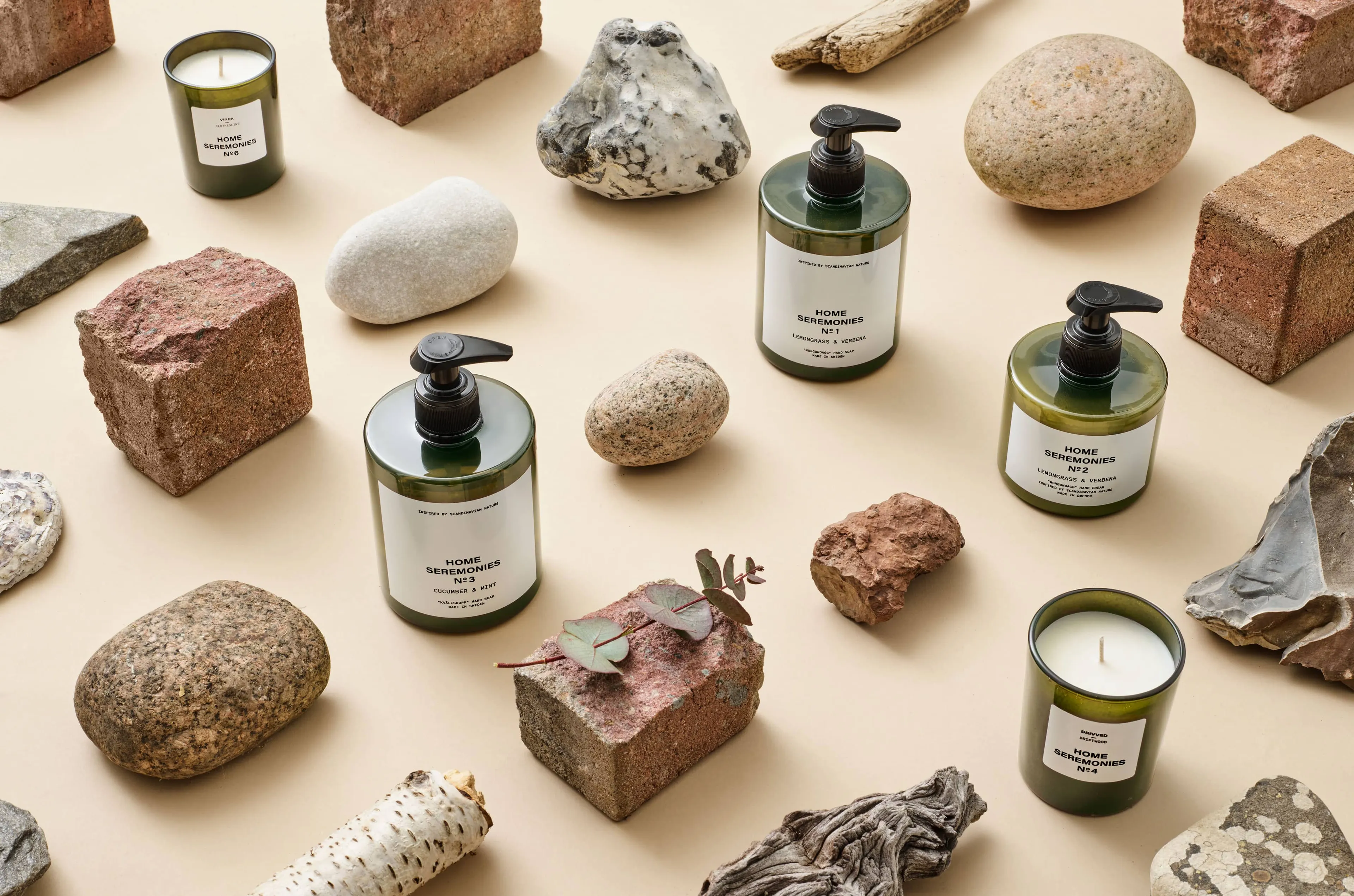 Fyra mörkgröna tvålpumpar och två gröna doftljus placerade bland stenar, träbitar och tegelstenar på en ljus yta. Produkterna har enkla vita etiketter med svart text.