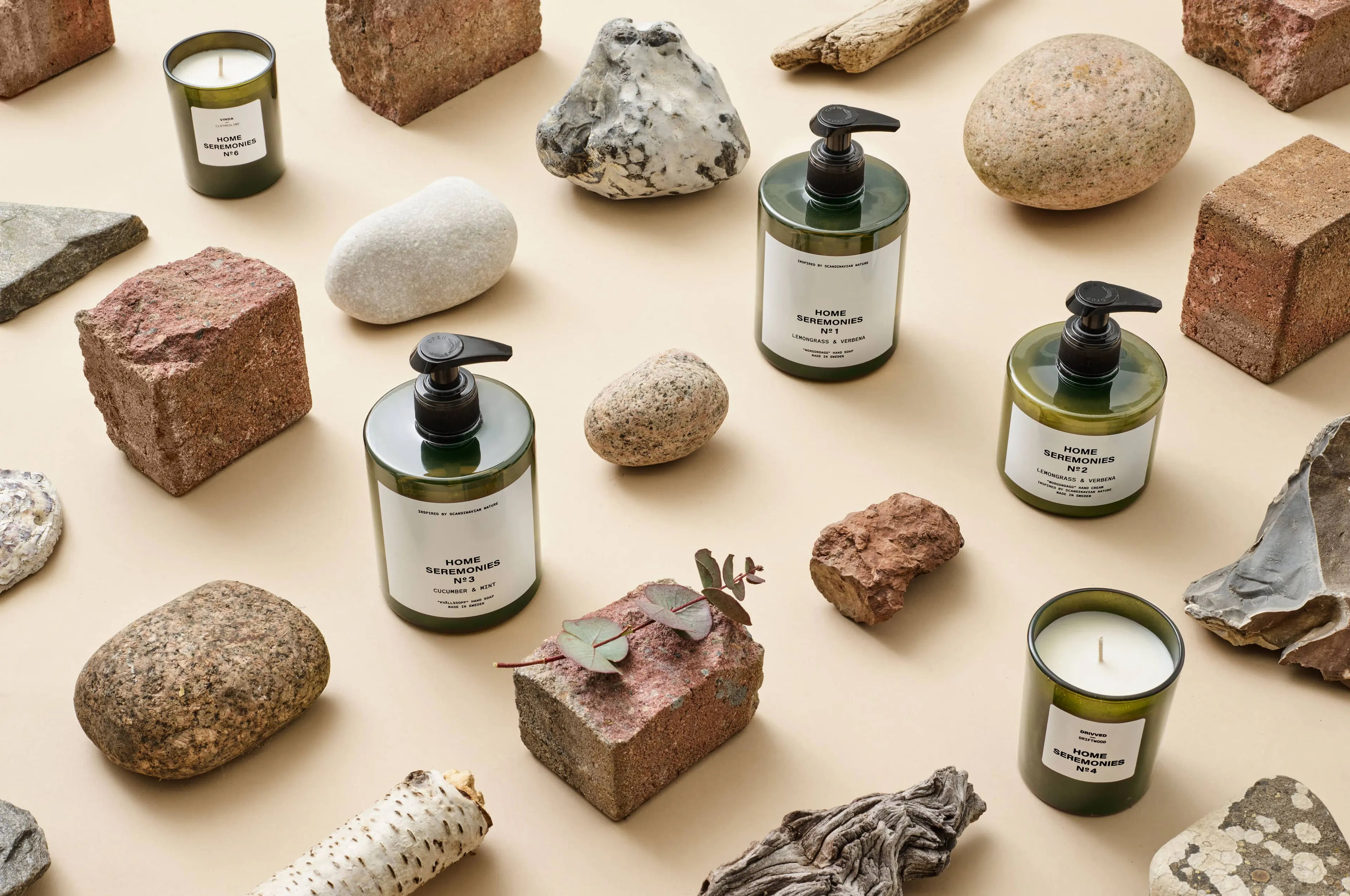 Fyra mörkgröna tvålpumpar och två gröna doftljus placerade bland stenar, träbitar och tegelstenar på en ljus yta. Produkterna har enkla vita etiketter med svart text.