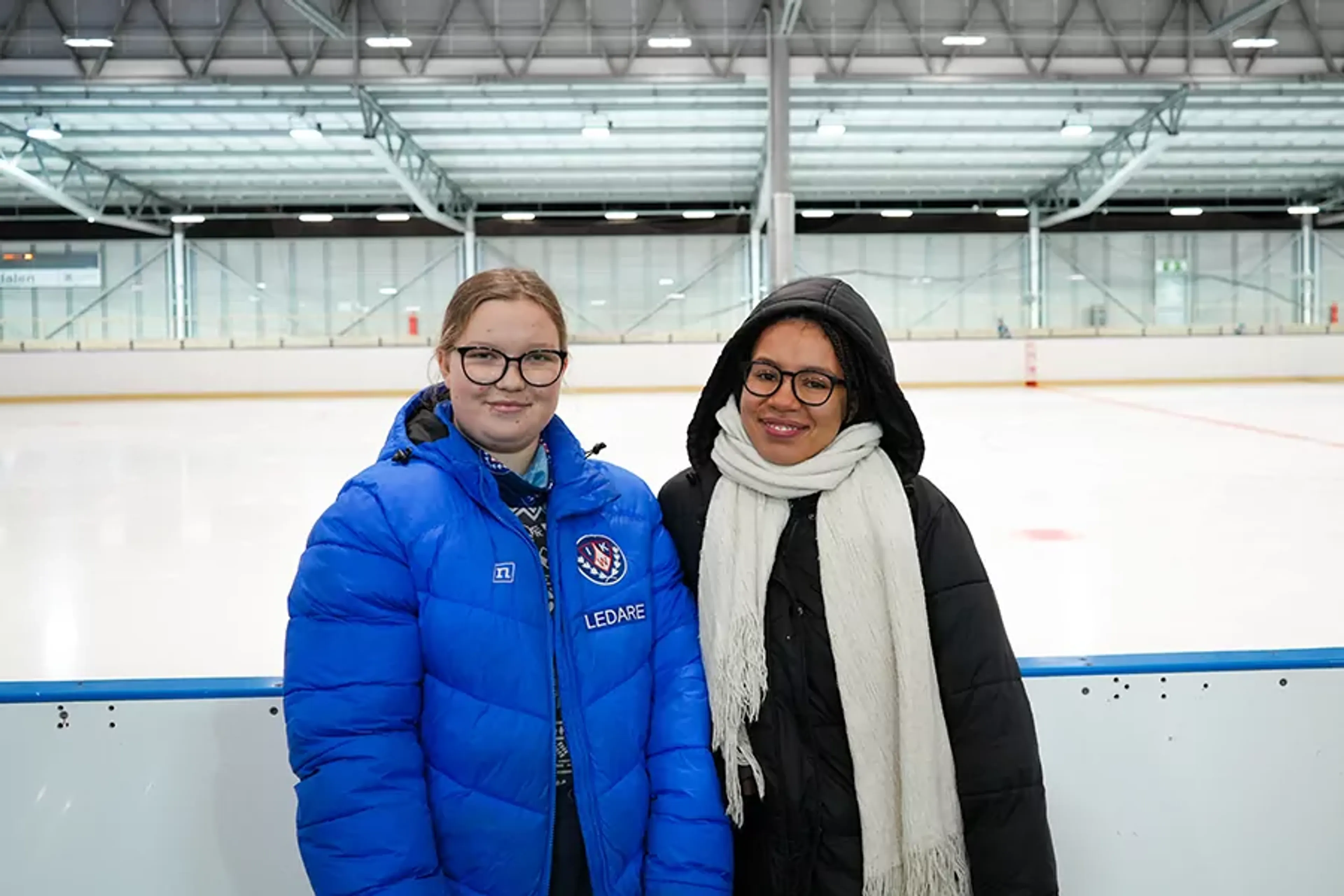 Två unga ledare står vid sidan av en tom ishall, klädda i varma jackor och glasögon. Den ena bär en blå jacka med texten "Ledare" på bröstet.