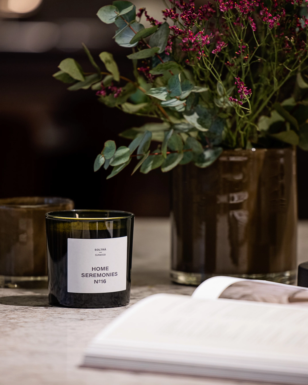 Ett doftljus i mörkt glas med texten "Home Seremonies" på en marmorbordsskiva, omgiven av en vas med gröna blad och lila blommor samt en öppen bok.