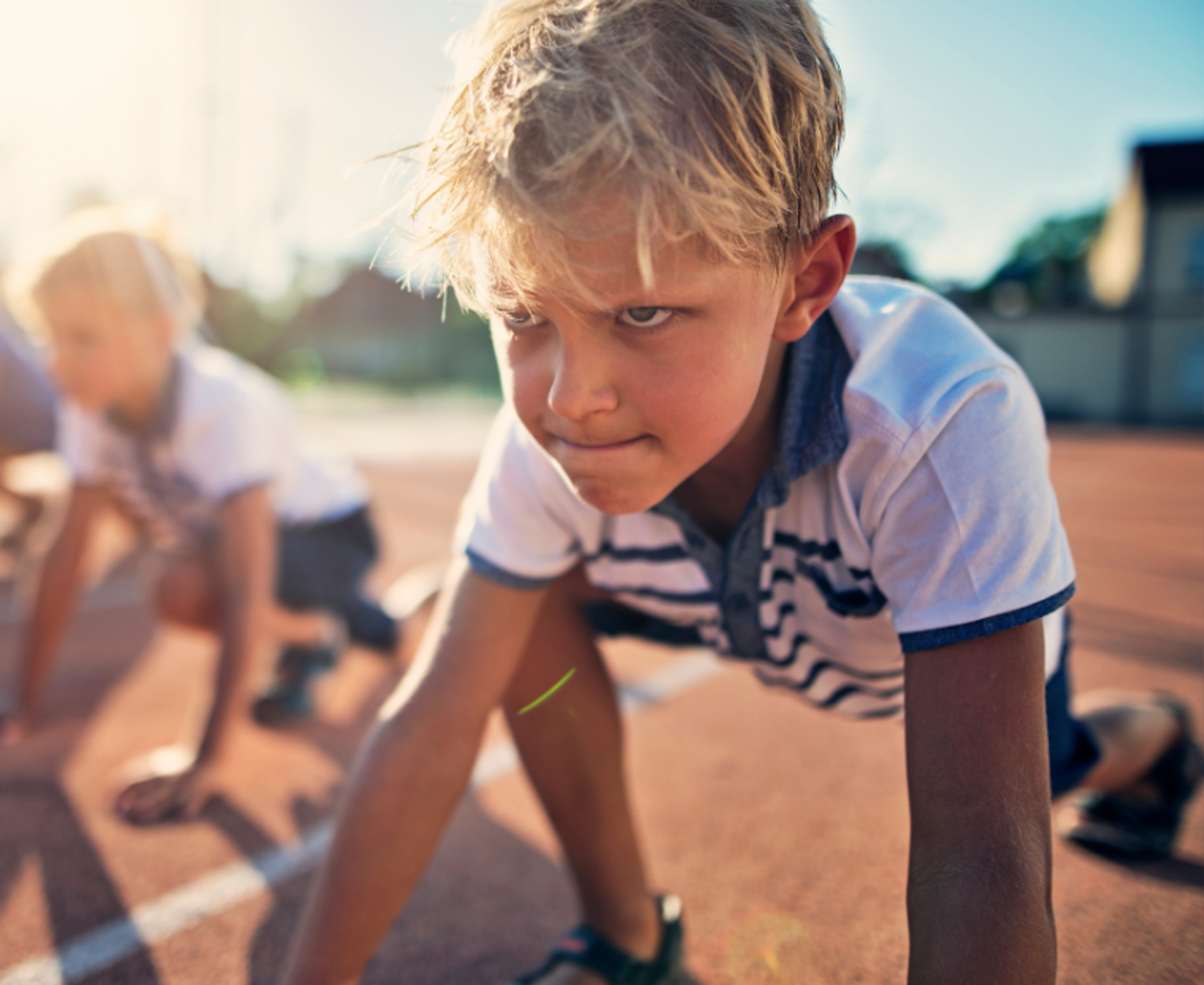 Tre barn i startblocken på en löparbana, redo att springa. Solen skapar långa skuggor över den röda underlaget.