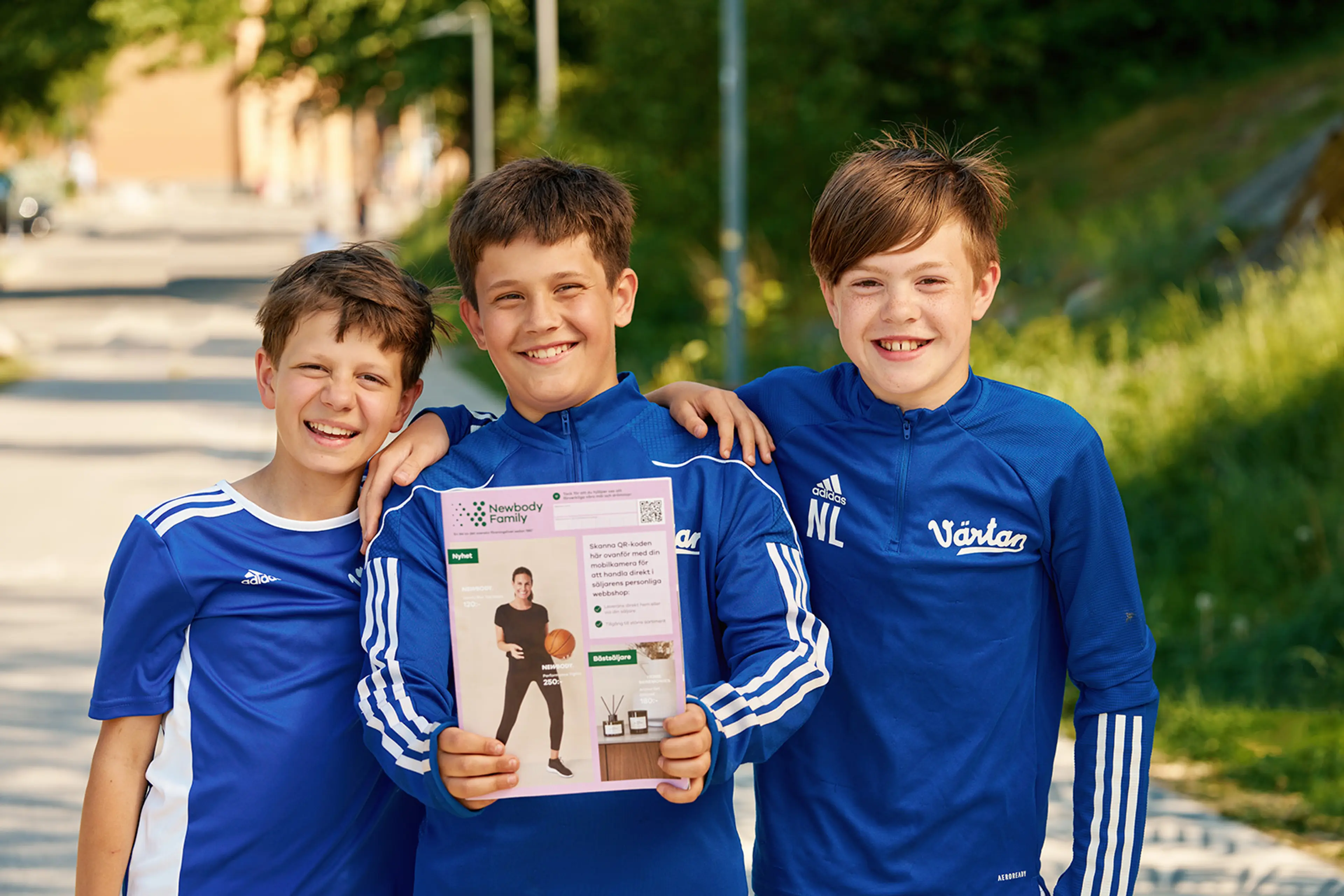 Tre glada pojkar i blå sportkläder står tillsammans utomhus, där mittenpojken håller upp en katalog. Katalogen har en ljusrosa framsida med bilder och text.