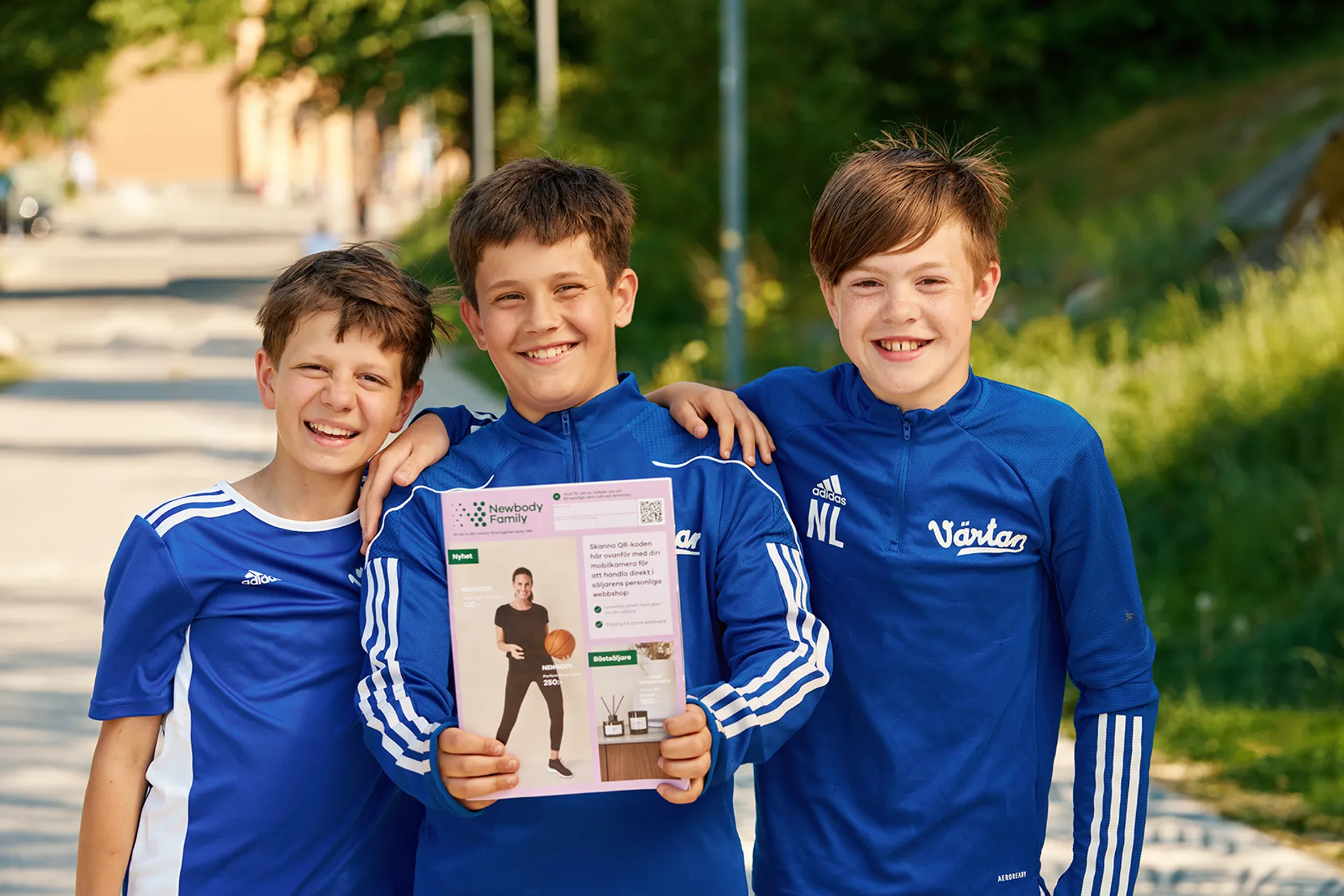 Tre glada pojkar i blå sportkläder står tillsammans utomhus, där mittenpojken håller upp en katalog. Katalogen har en ljusrosa framsida med bilder och text.