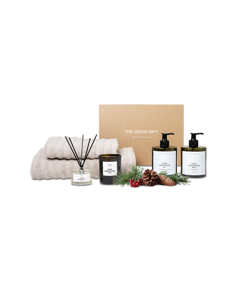 Ett set med två beige handdukar, en doftljus, doftpinnar och två pumpflaskor med tvål eller lotion placerade framför en presentkartong. En kott- och bärdekoration ligger som accent bredvid produkterna.