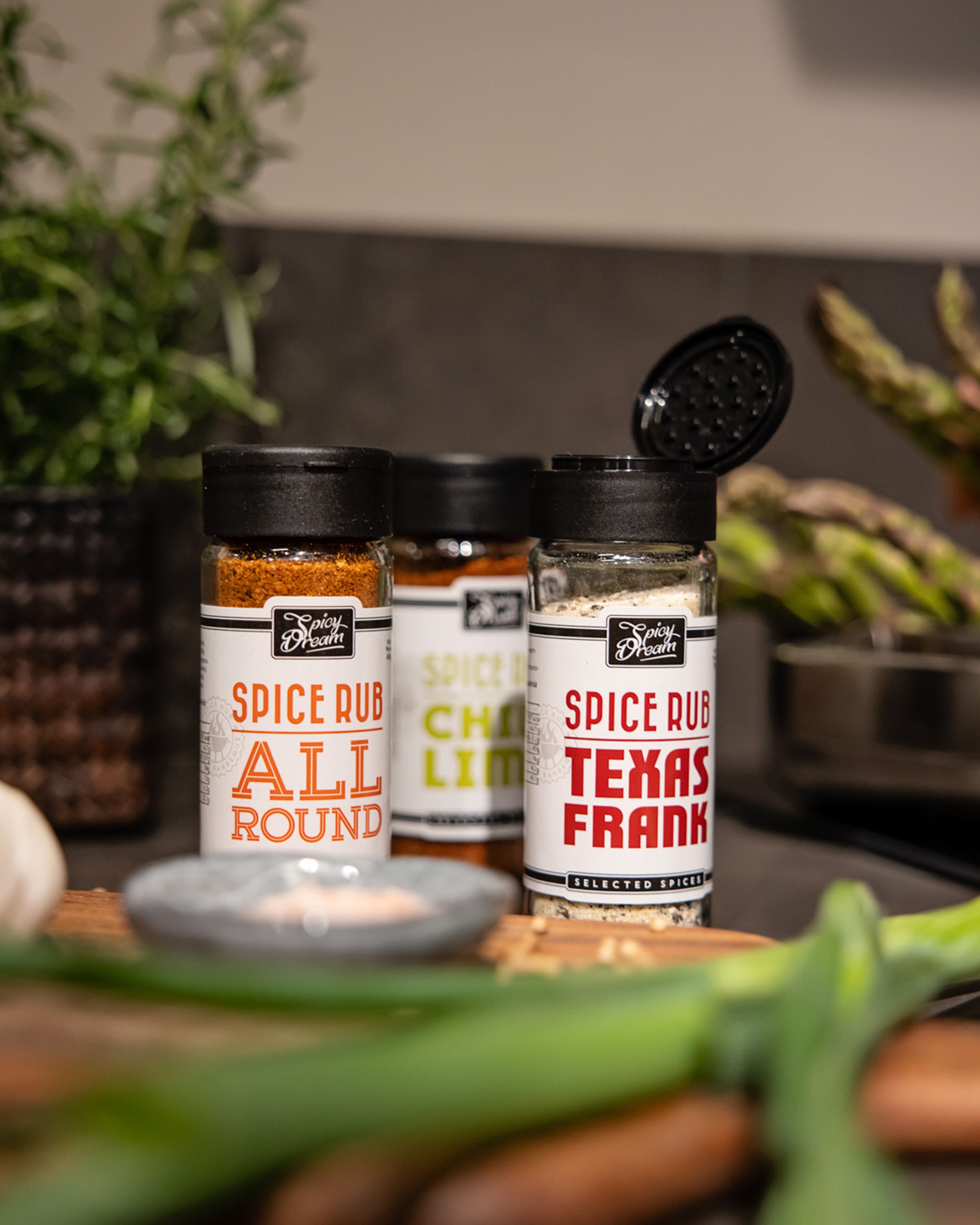 Tre kryddburkar med olika spice rubs, varav en med öppet lock visar ett ljust kryddpulver. Färska grönsaker och örter syns i förgrunden och bakgrunden.