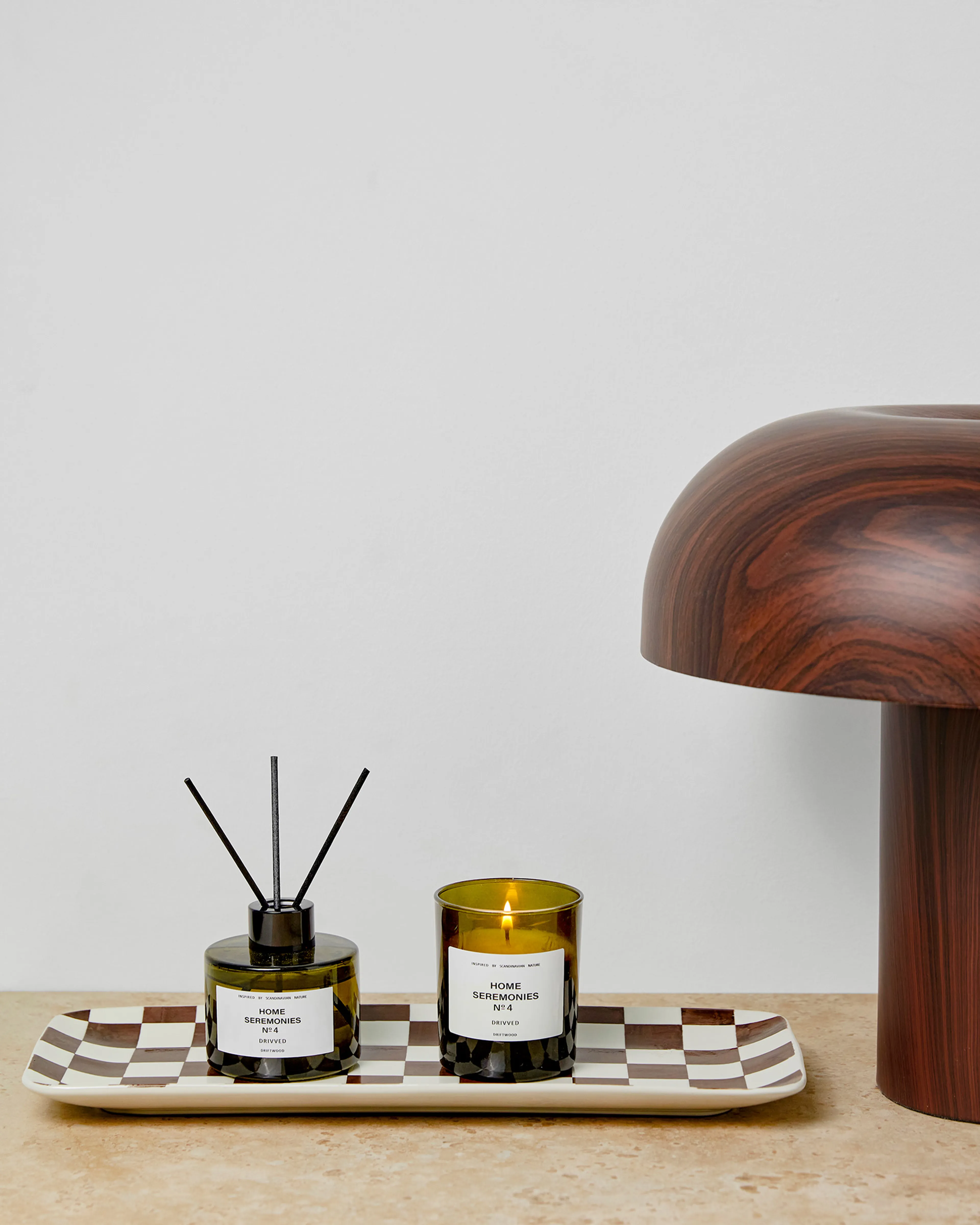 Doftset med doftspridare och tända doftljus i grönton på ett rutigt fat, placerade på en yta bredvid en brun lampskärm i träimitation.