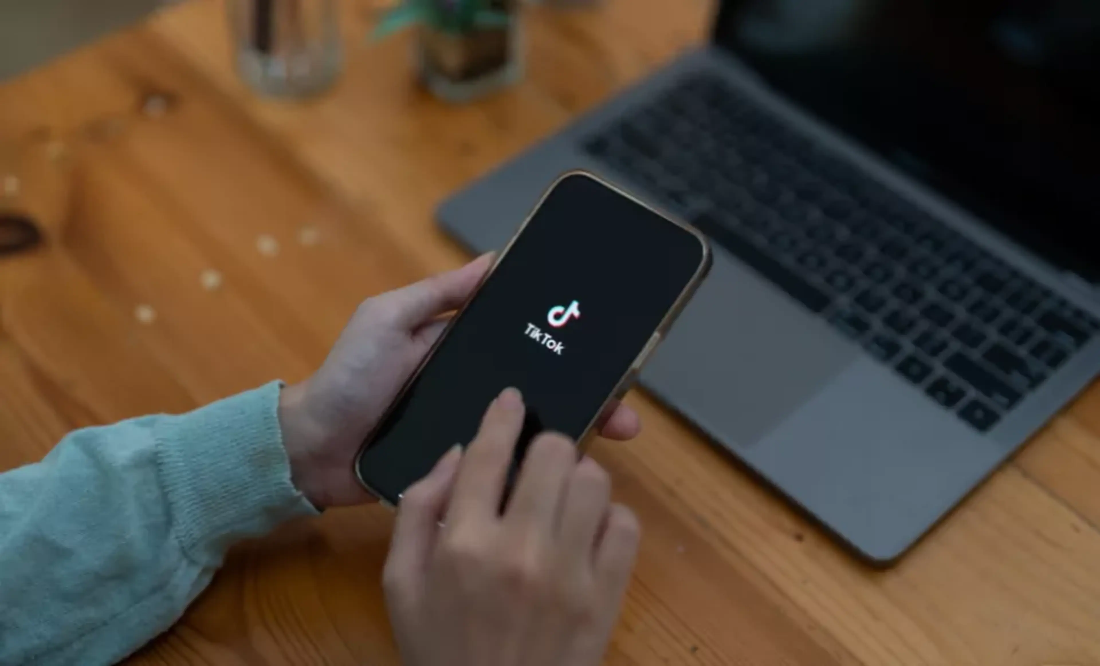 En person håller en mobiltelefon med TikTok-appen öppen. En öppen laptop står på ett träbord i bakgrunden.