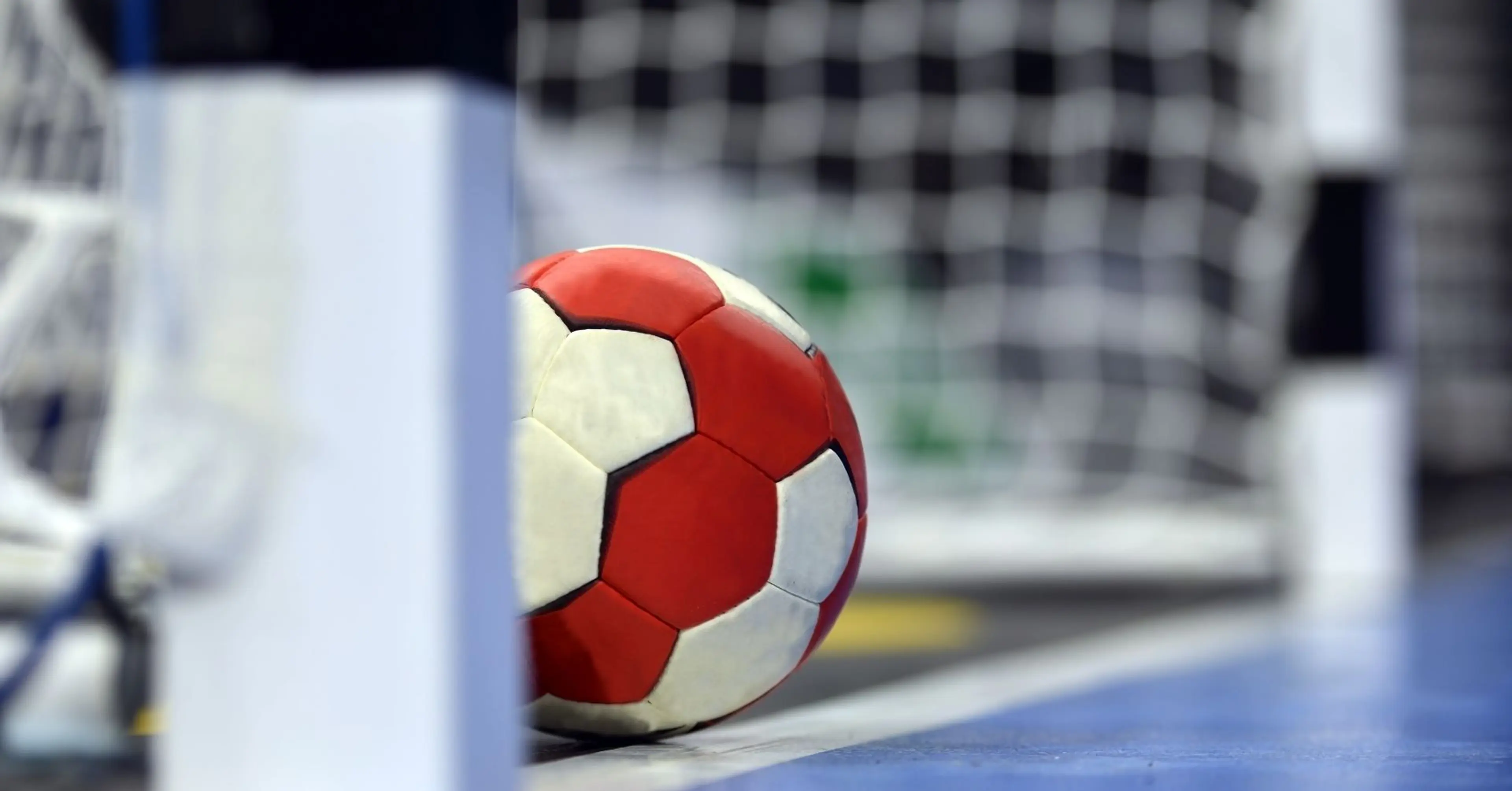 En röd- och vitmönstrad handboll som ligger vid mållinjen framför ett handbollsmål. Golvet är blått och bakgrunden suddig med nätet tydligt synligt.