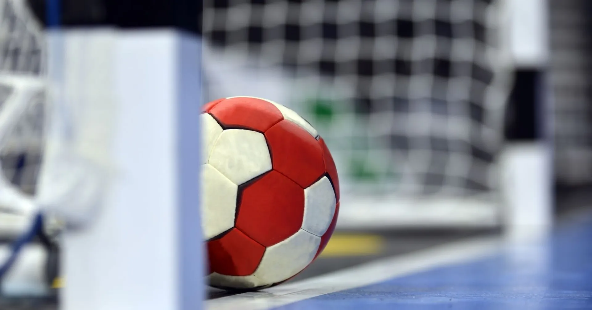 En röd- och vitmönstrad handboll som ligger vid mållinjen framför ett handbollsmål. Golvet är blått och bakgrunden suddig med nätet tydligt synligt.