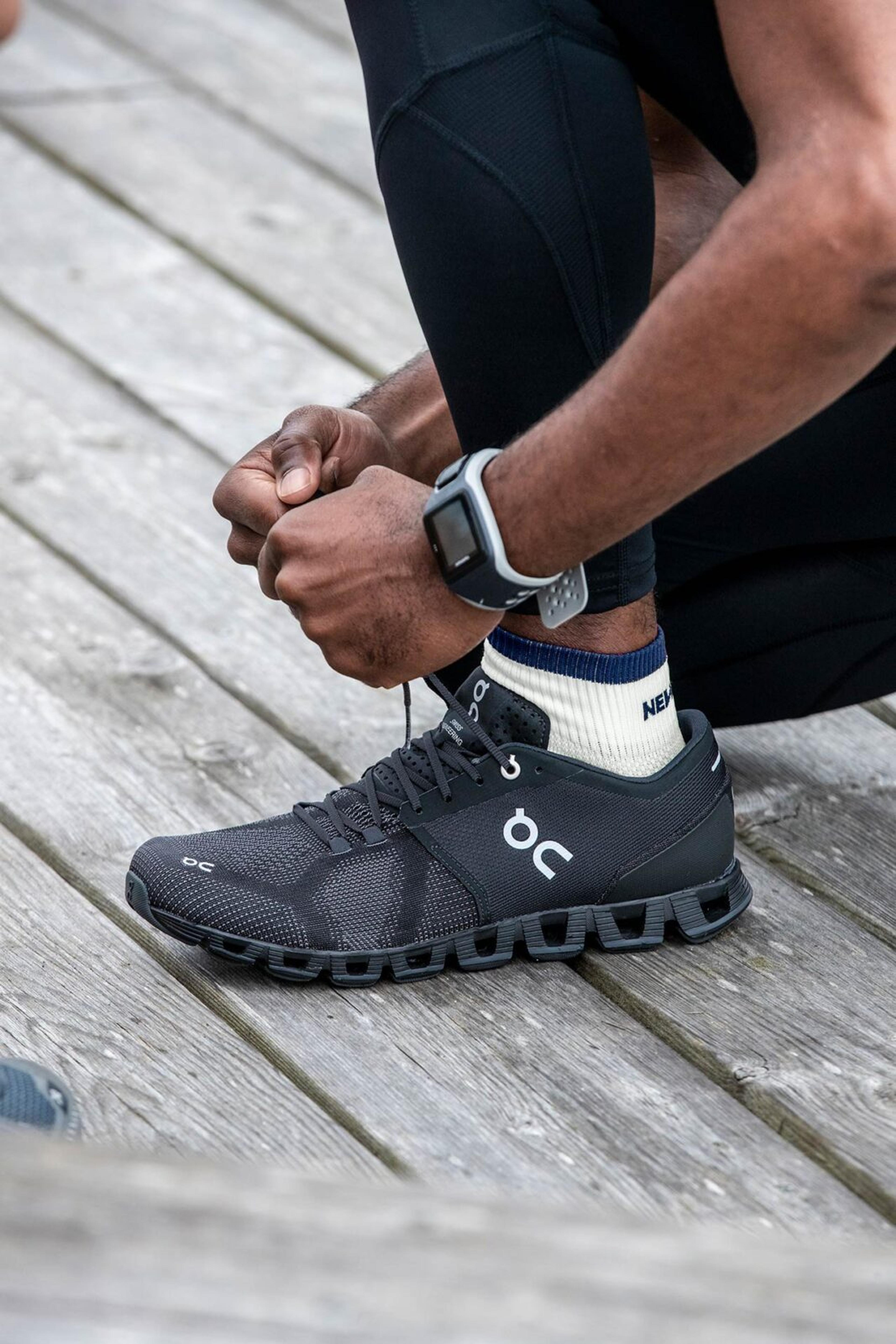En person som knyter skosnöret på en svart löparsko med vita detaljer, iklädd vita strumpor med mörkblå kant och sportiga träningskläder.