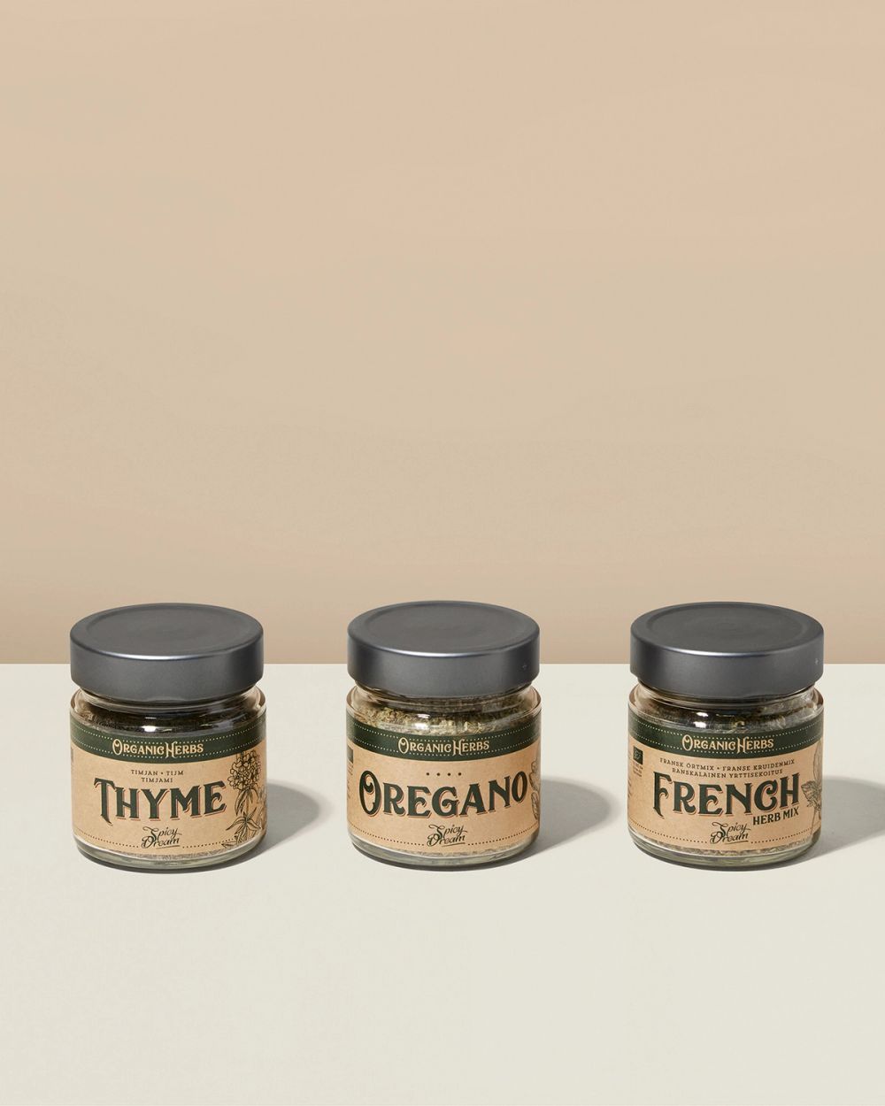 Tre glasburkar med ekologiska kryddor, timjan, oregano och en fransk örtkryddblandning, med beige etiketter och mörka lock.