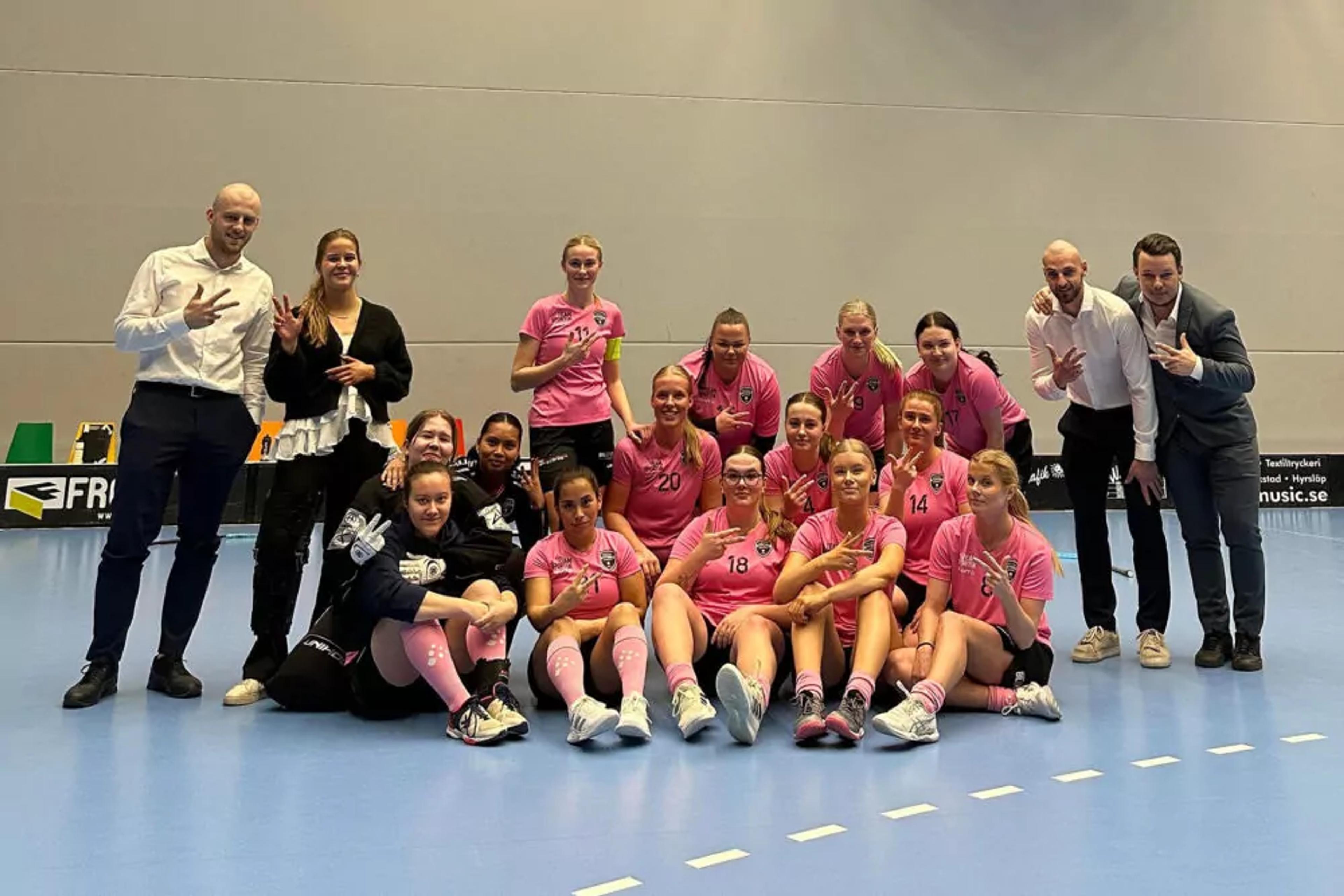 En handbollstrupp med spelare i rosa matchtröjor och strumpor, omgiven av ledare i vardagskläder på en inomhusplan. Gruppen sitter och står samlad, alla visar tecken med händerna.