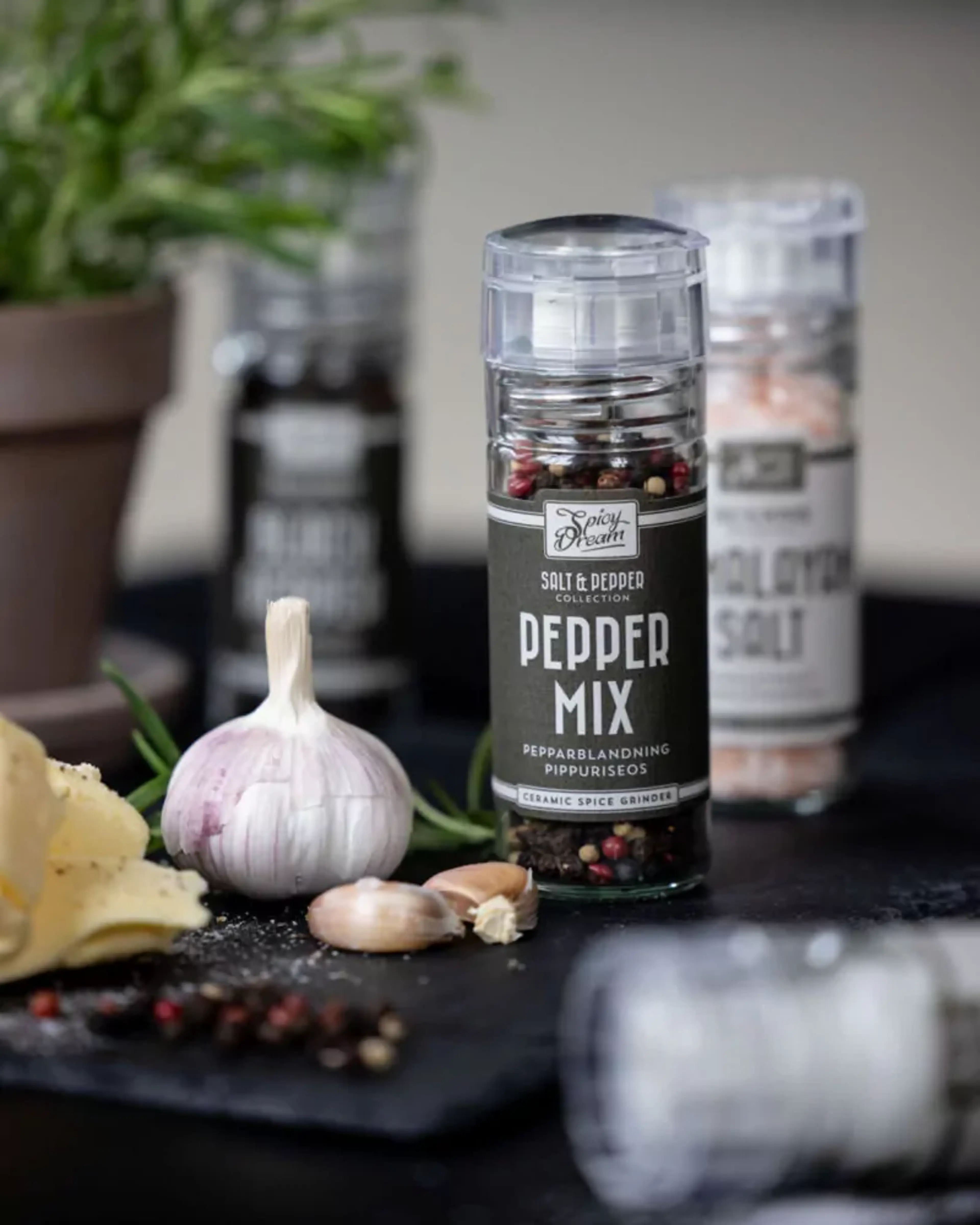 En förpackning med mixad peppar i en genomskinlig kvarn med svart etikett, omgiven av vitlök och ost. Bakom syns salt och svartpeppar i liknande kvarnar.