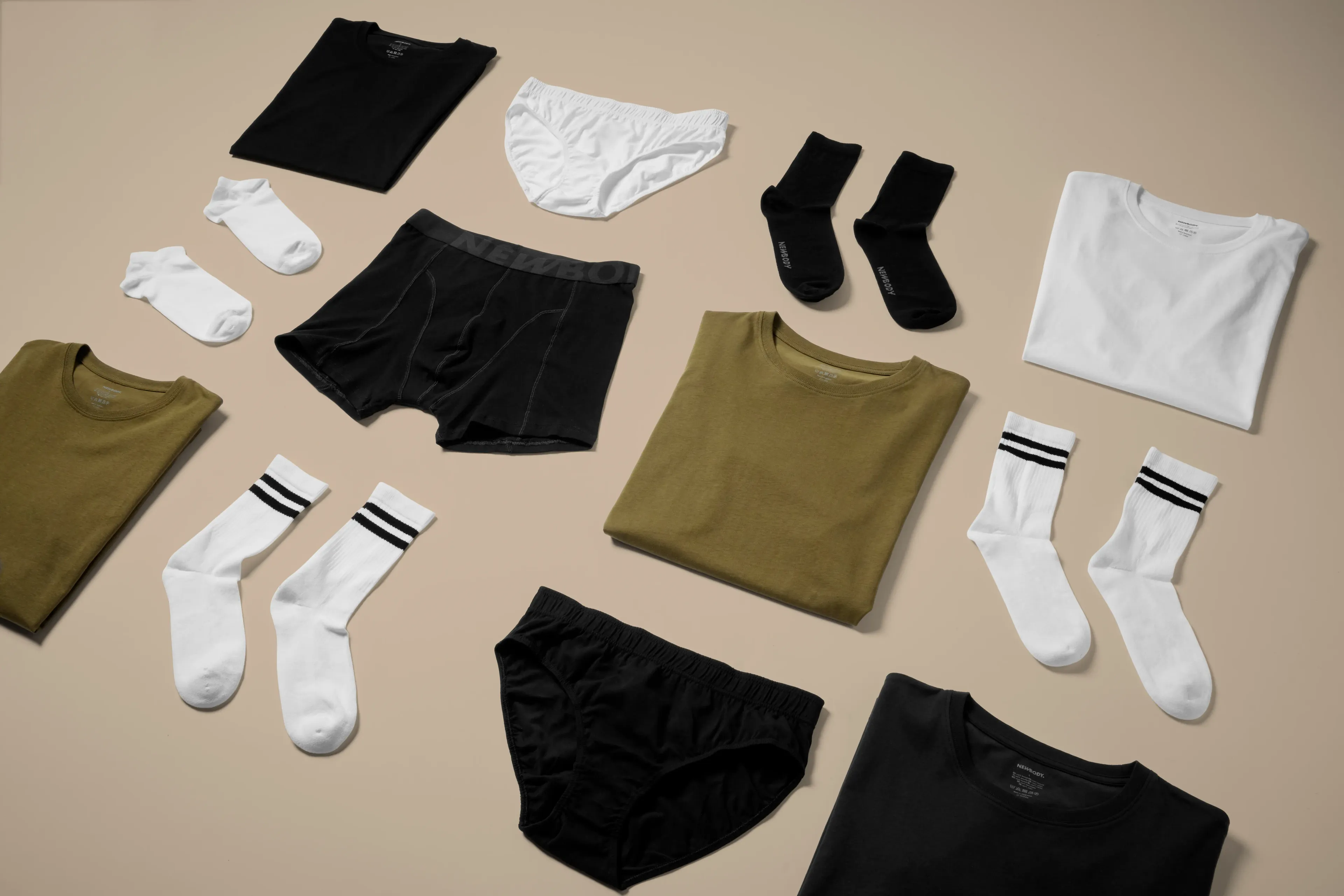 Olika plagg i neutrala färger, bland annat t-shirts, sockor och underkläder, snyggt upplagda på ett ljust underlag. Enkel design med fokus på basplagg för alla tillfällen.