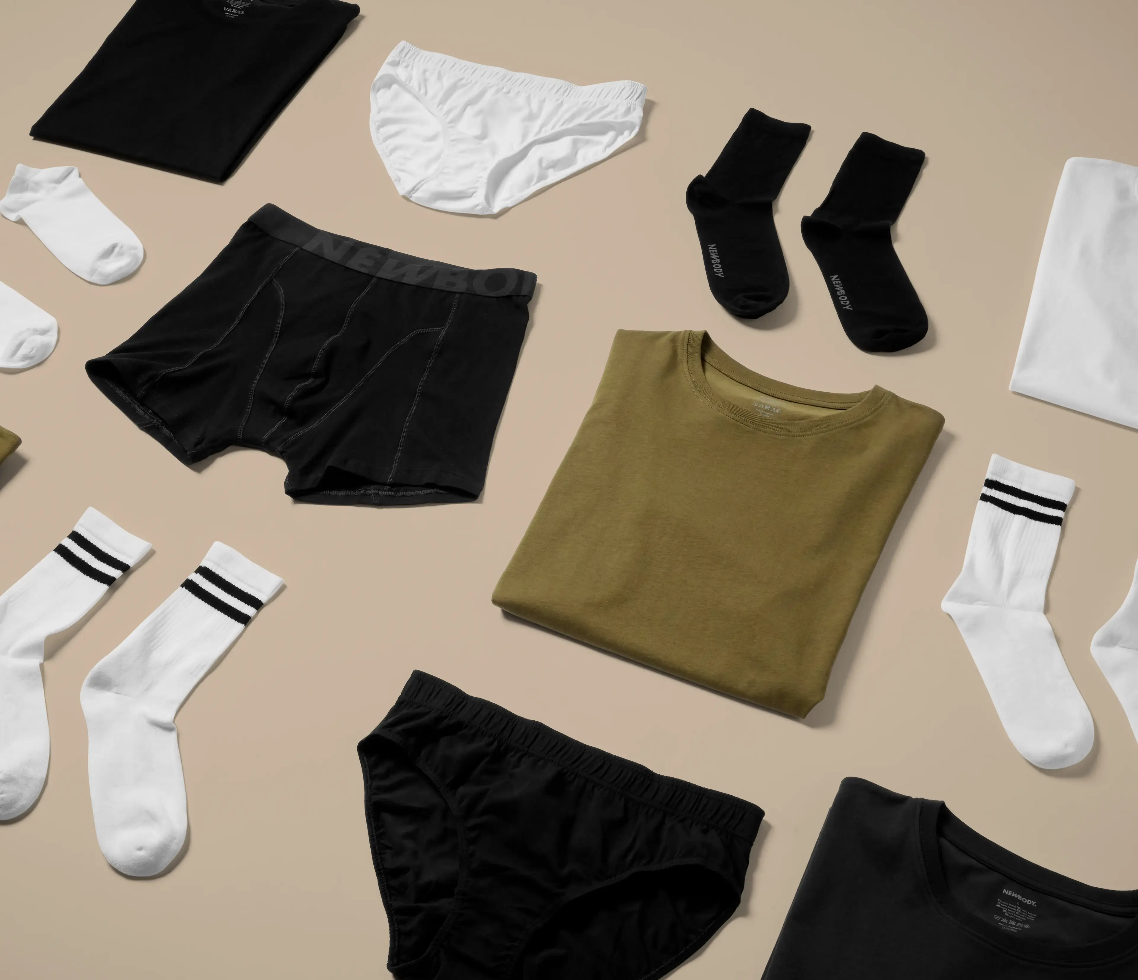 Olika plagg i neutrala färger, bland annat t-shirts, sockor och underkläder, snyggt upplagda på ett ljust underlag. Enkel design med fokus på basplagg för alla tillfällen.