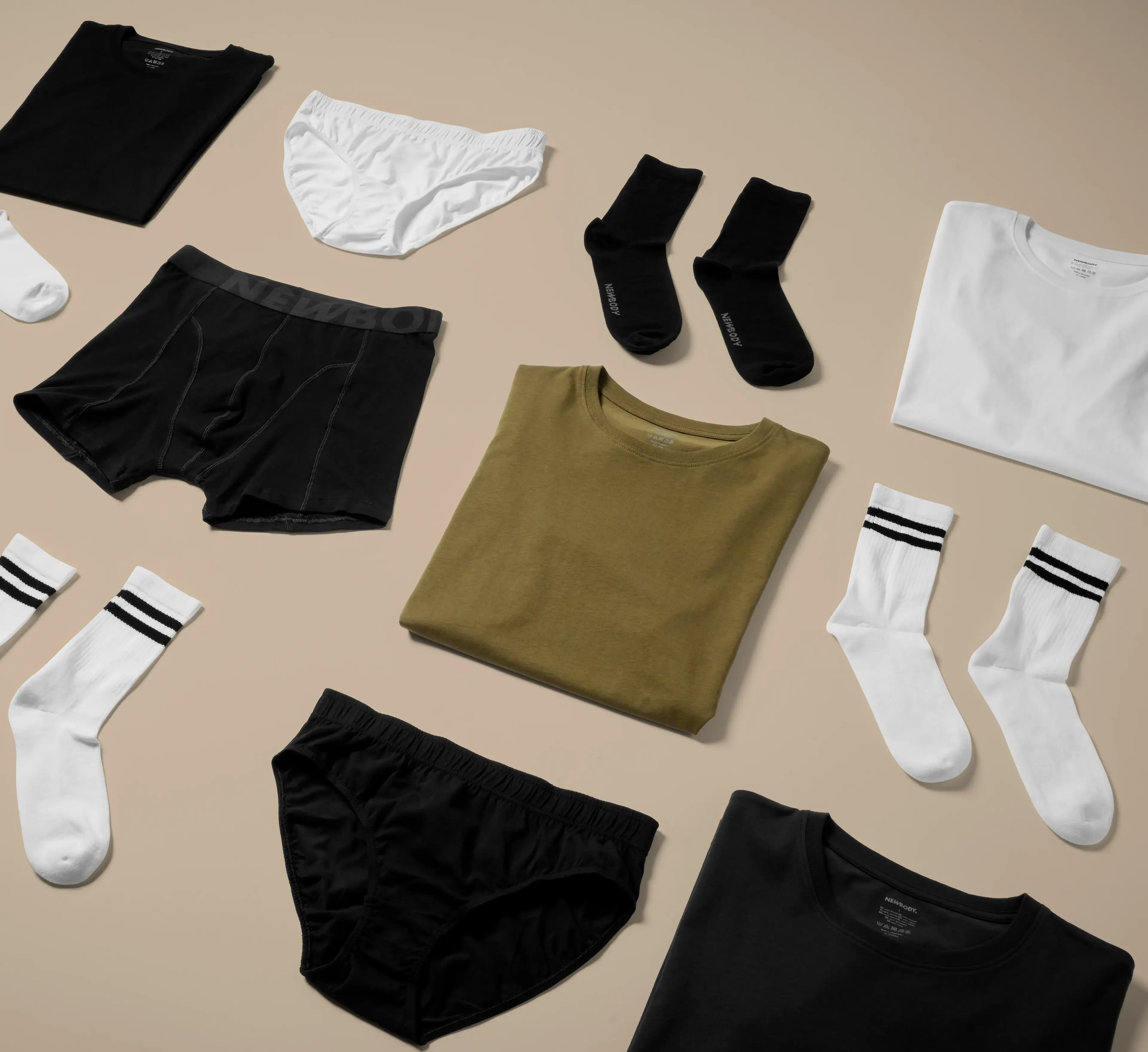 Olika plagg i neutrala färger, bland annat t-shirts, sockor och underkläder, snyggt upplagda på ett ljust underlag. Enkel design med fokus på basplagg för alla tillfällen.