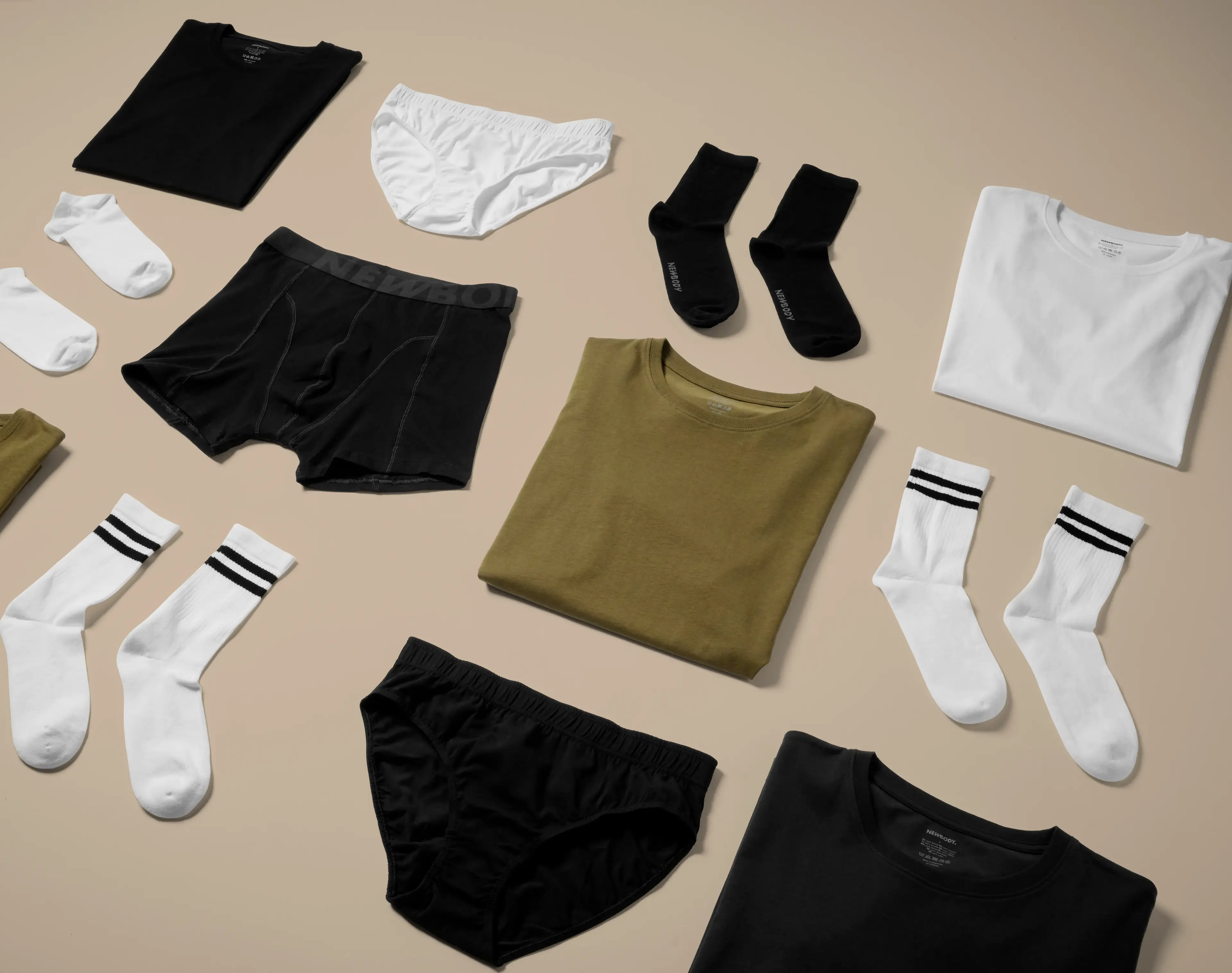 Olika plagg i neutrala färger, bland annat t-shirts, sockor och underkläder, snyggt upplagda på ett ljust underlag. Enkel design med fokus på basplagg för alla tillfällen.